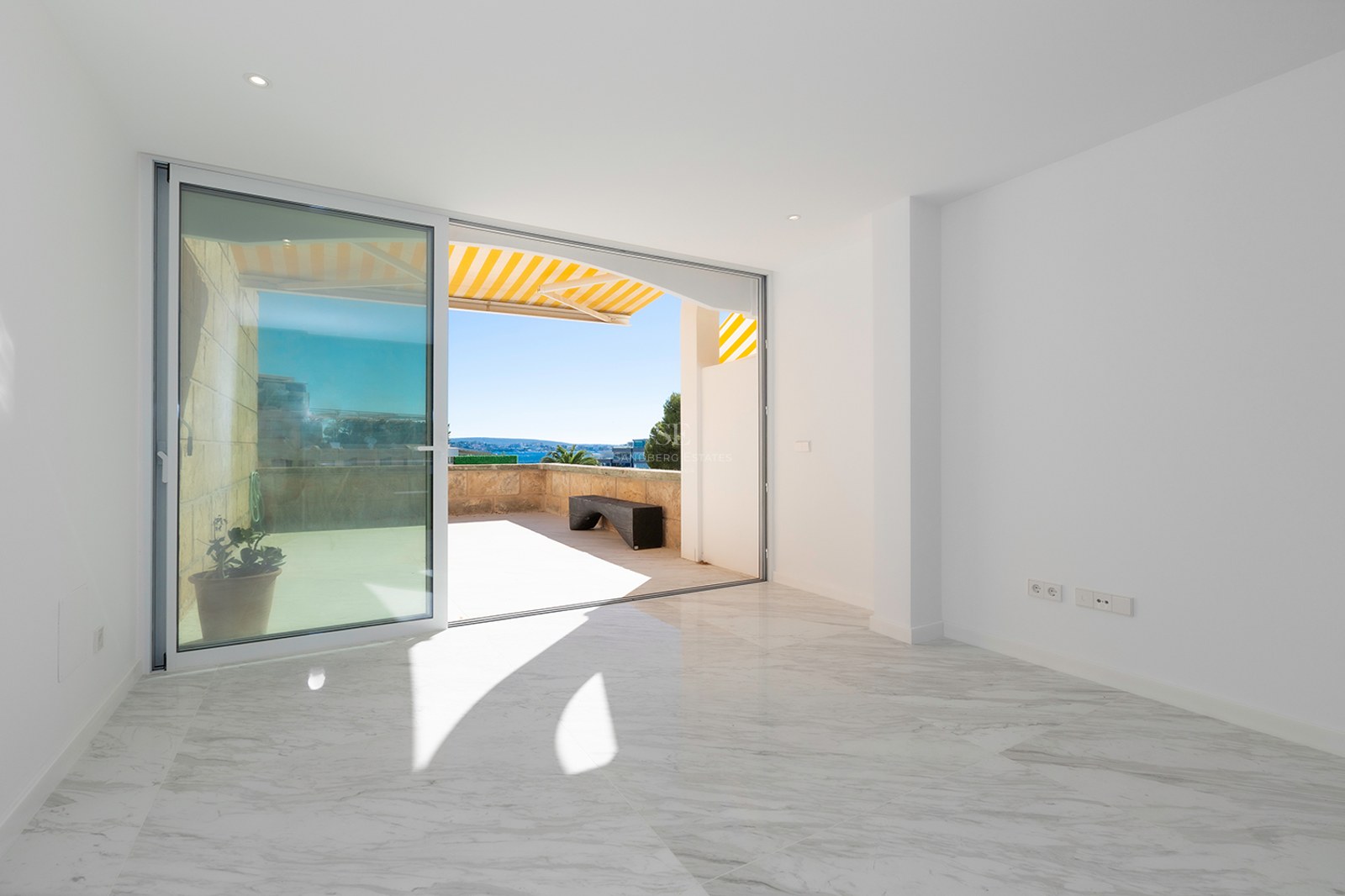 Bright empty room with white marble floors and a large sliding glass door opening to a sunny stone terrace.