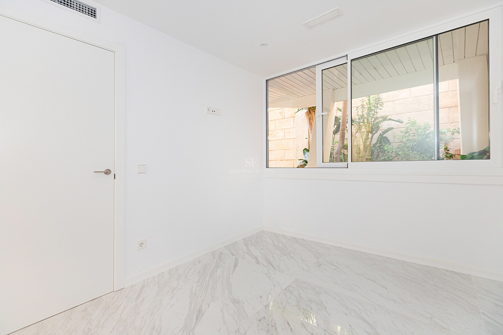 Empty room with white walls, polished marble floor, and a large sliding window overlooking outdoor greenery.