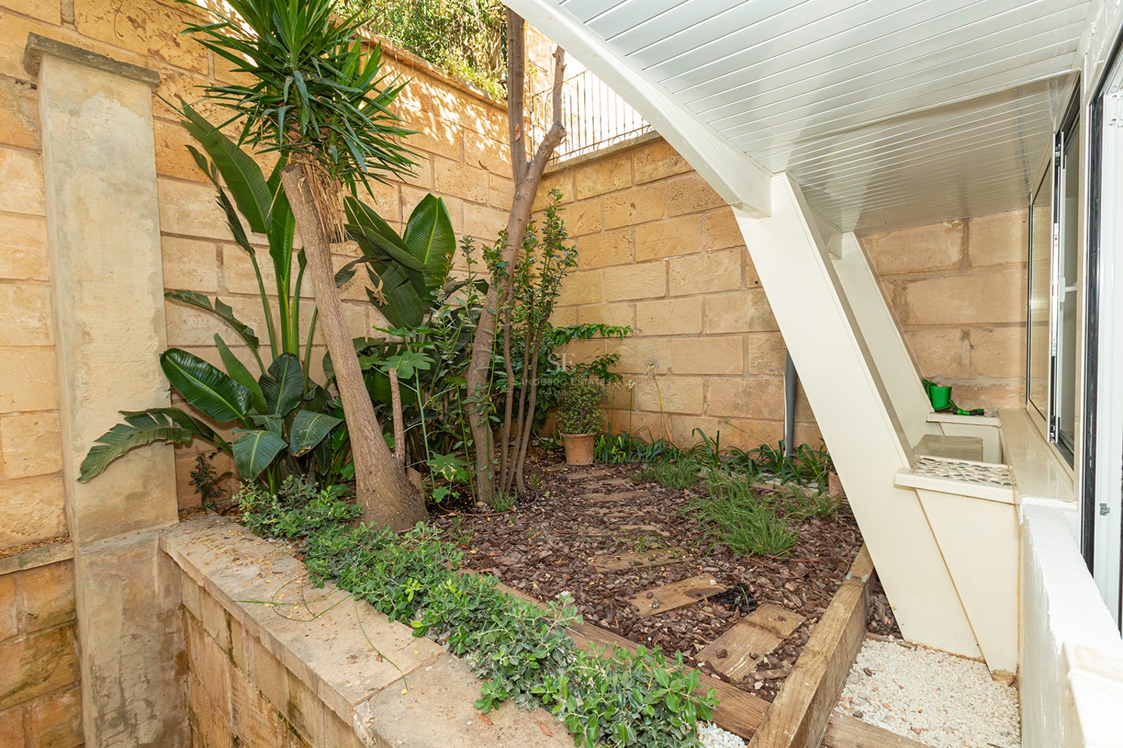 A small outdoor garden area with tropical plants, wood mulch, and a sandstone wall under a slanted white structure.