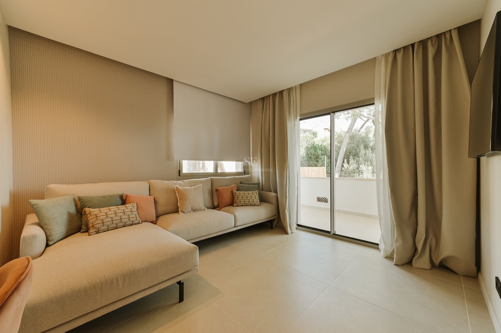 Salón moderno con sofá rinconera neutro, cojines en tonos tierra y grandes ventanales correderos hacia una terraza.