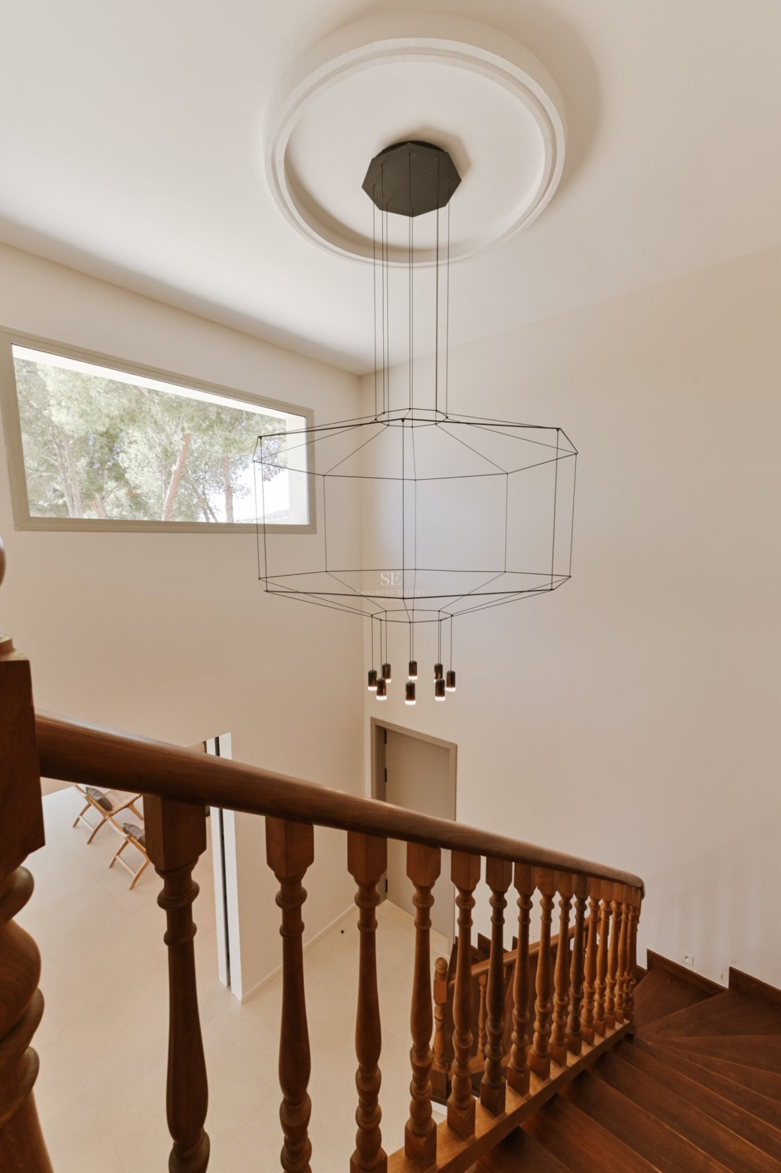 Barandilla de madera sobre vestíbulo de doble altura con lámpara geométrica de diseño y ventanal superior.