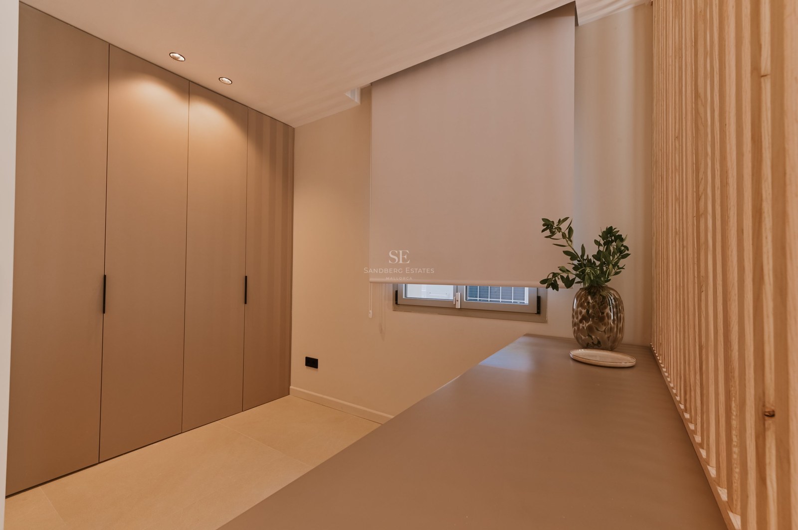 Dormitorio minimalista con armarios empotrados color topo, ventana con estor blanco y una encimera elegante.