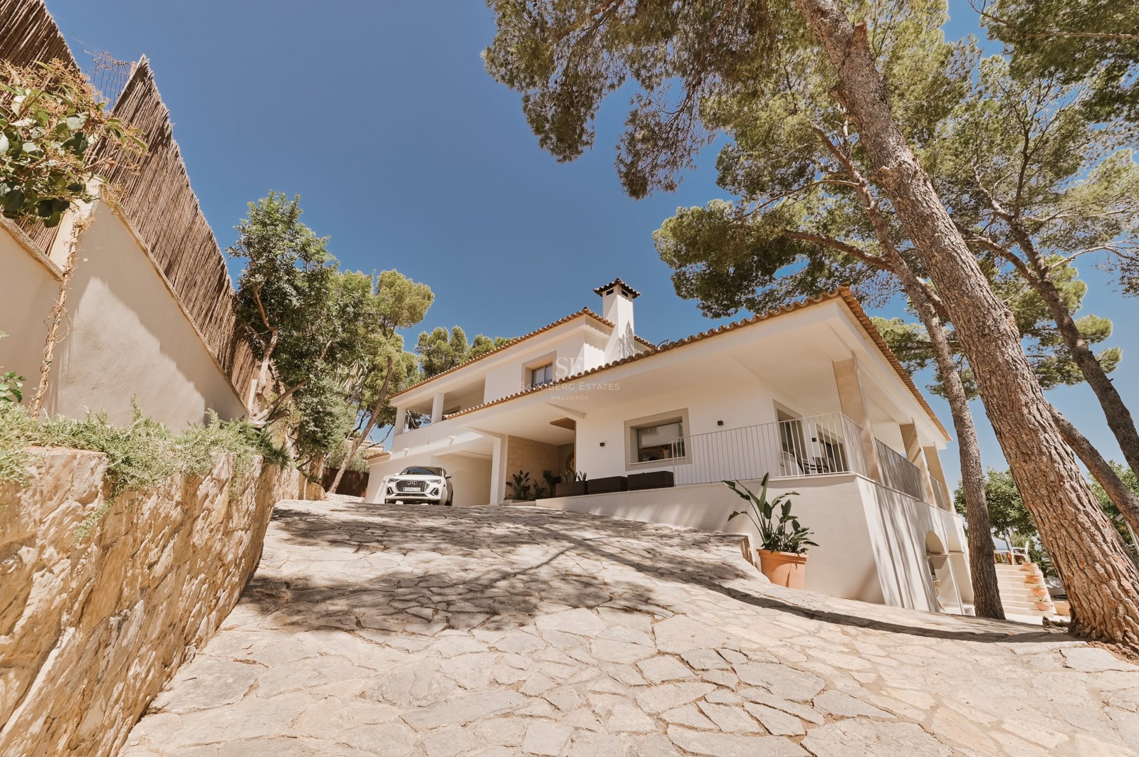 Vista exterior de una villa blanca de estilo mediterráneo con camino de piedra natural y pinos maduros.