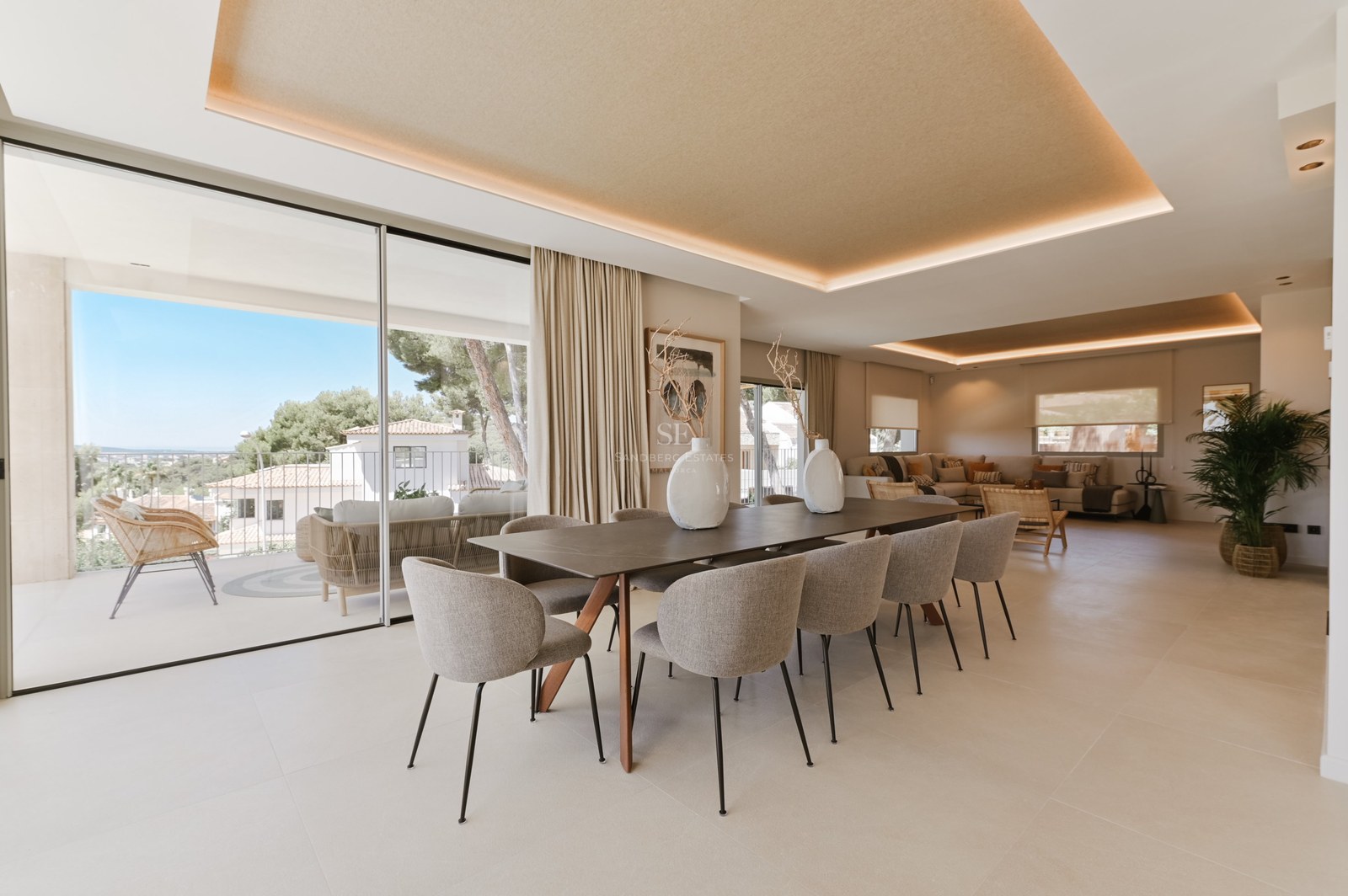 Comedor minimalista con mesa de madera, diez sillas de terciopelo gris y grandes ventanales correderos de cristal.