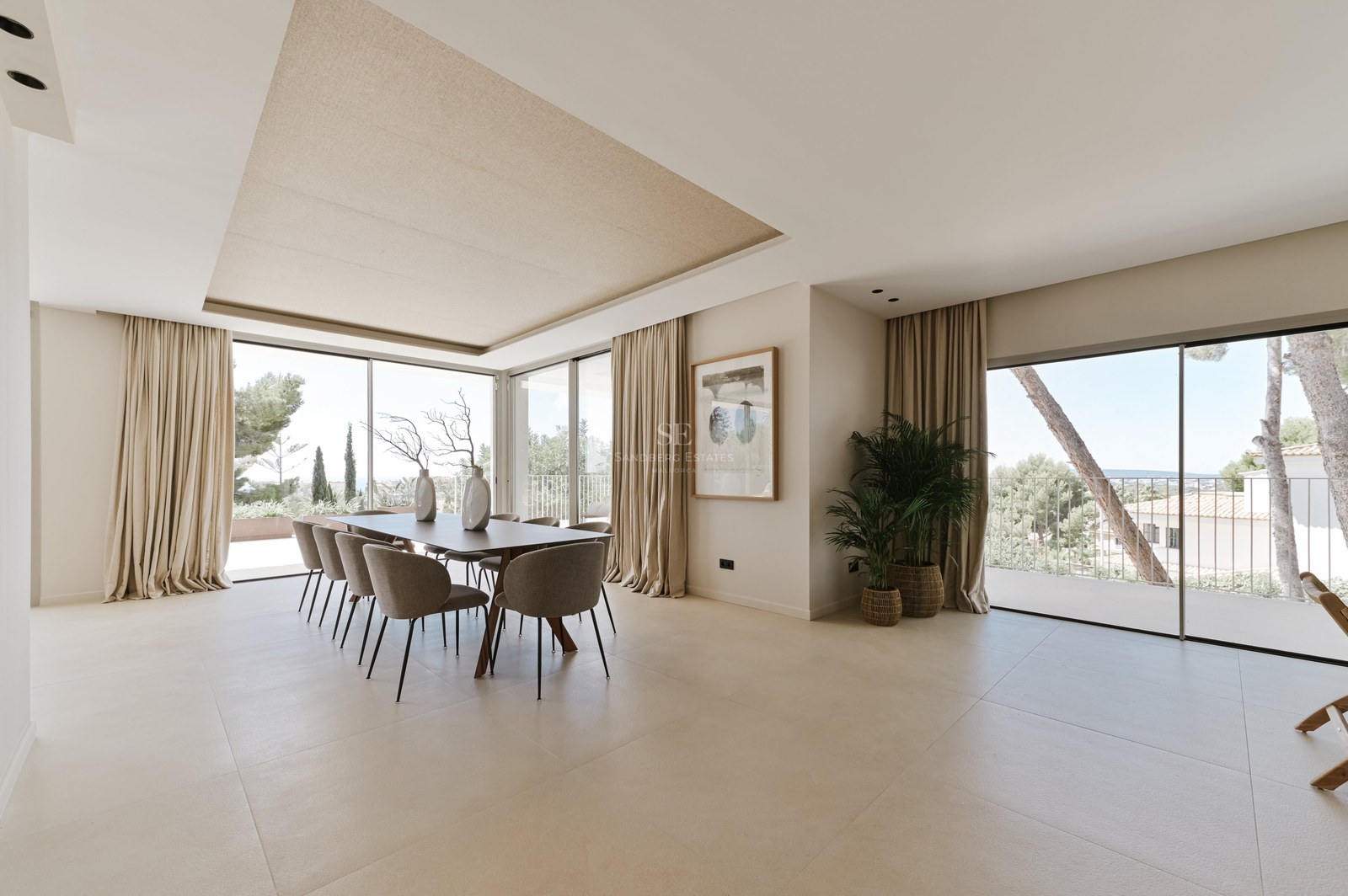 Comedor amplio con mesa para ocho, ventanales de suelo a techo, suelos de piedra clara y luz natural abundante.