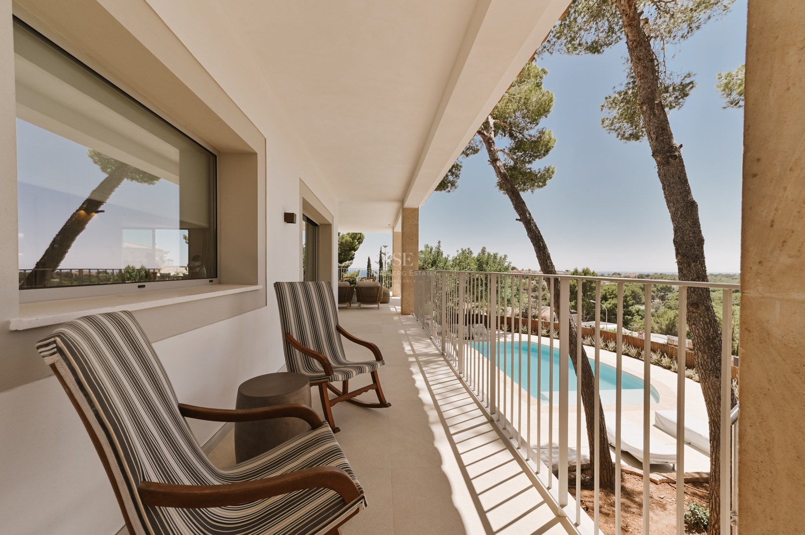 Terraza blanca cubierta con dos mecedoras de rayas con vistas a una piscina privada y pinos mediterráneos.