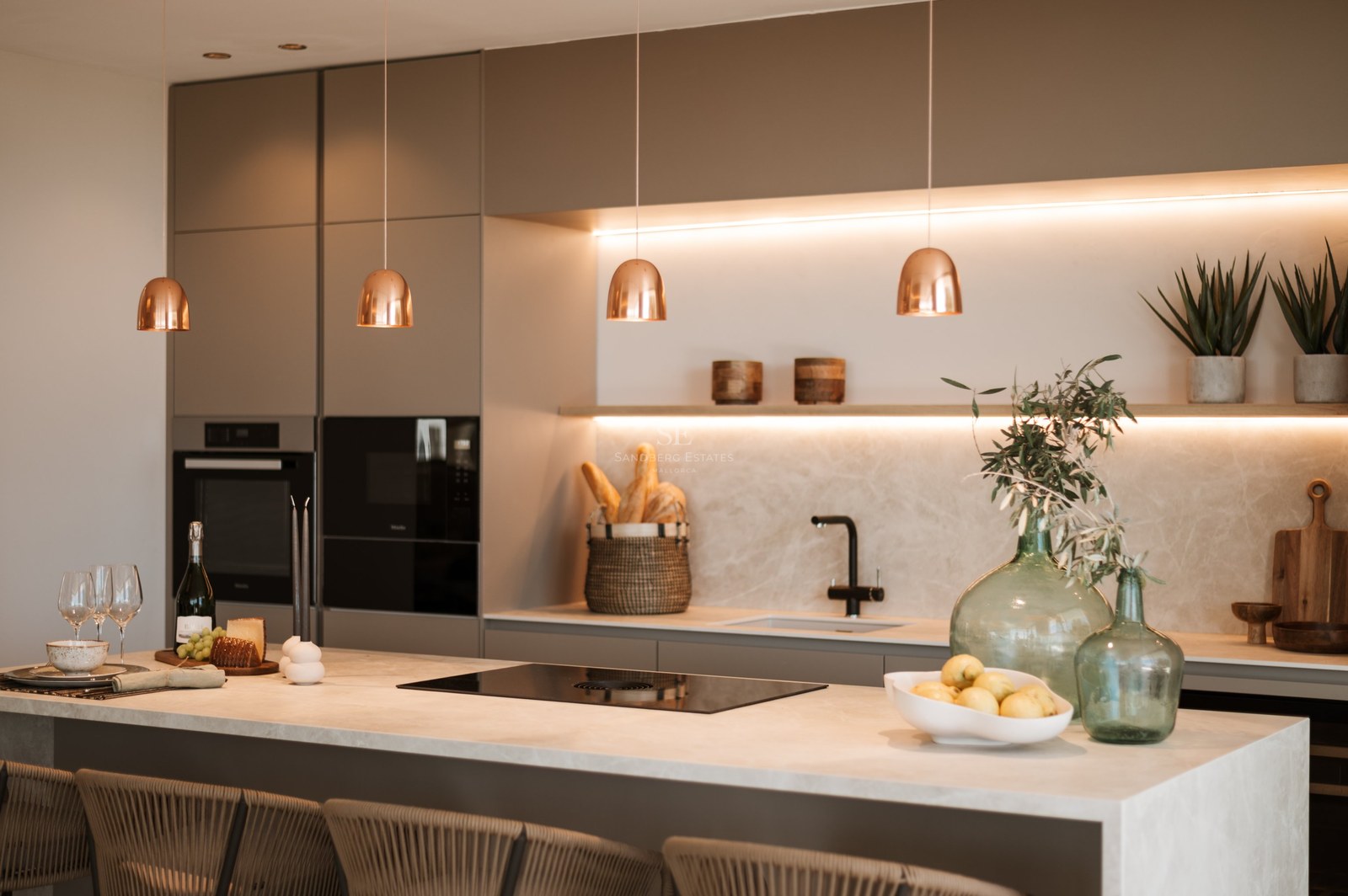 Cocina moderna con armarios color topo, salpicadero de mármol, lámparas de cobre e isla con taburetes.
