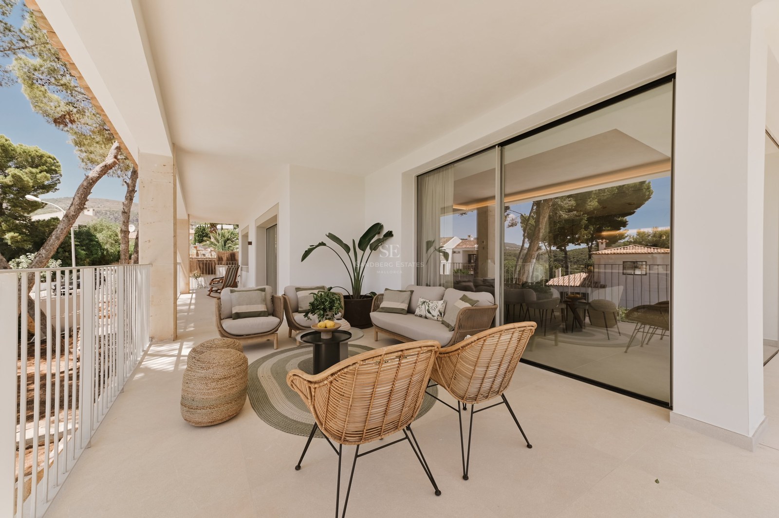 Terraza cubierta moderna con sillones de mimbre, sofá beige y amplios ventanales correderos que reflejan el exterior.