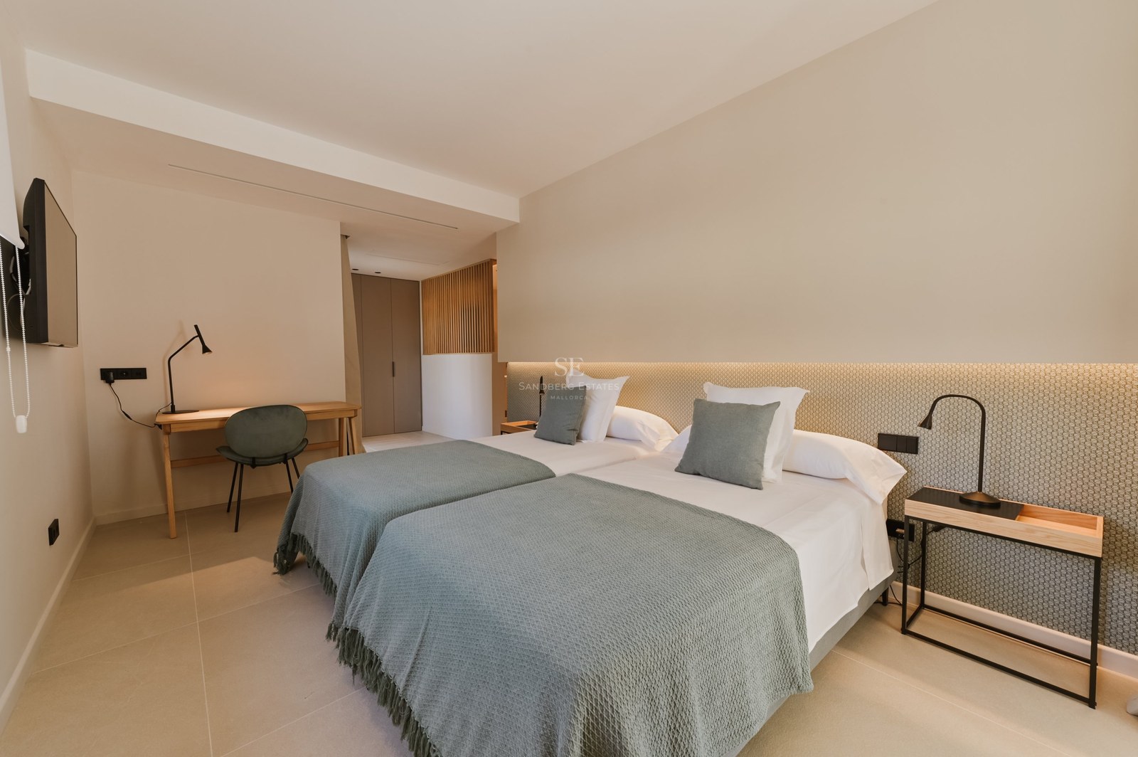 Dormitorio moderno con dos camas individuales, mantas verde salvia, escritorio de madera y luz cálida indirecta.