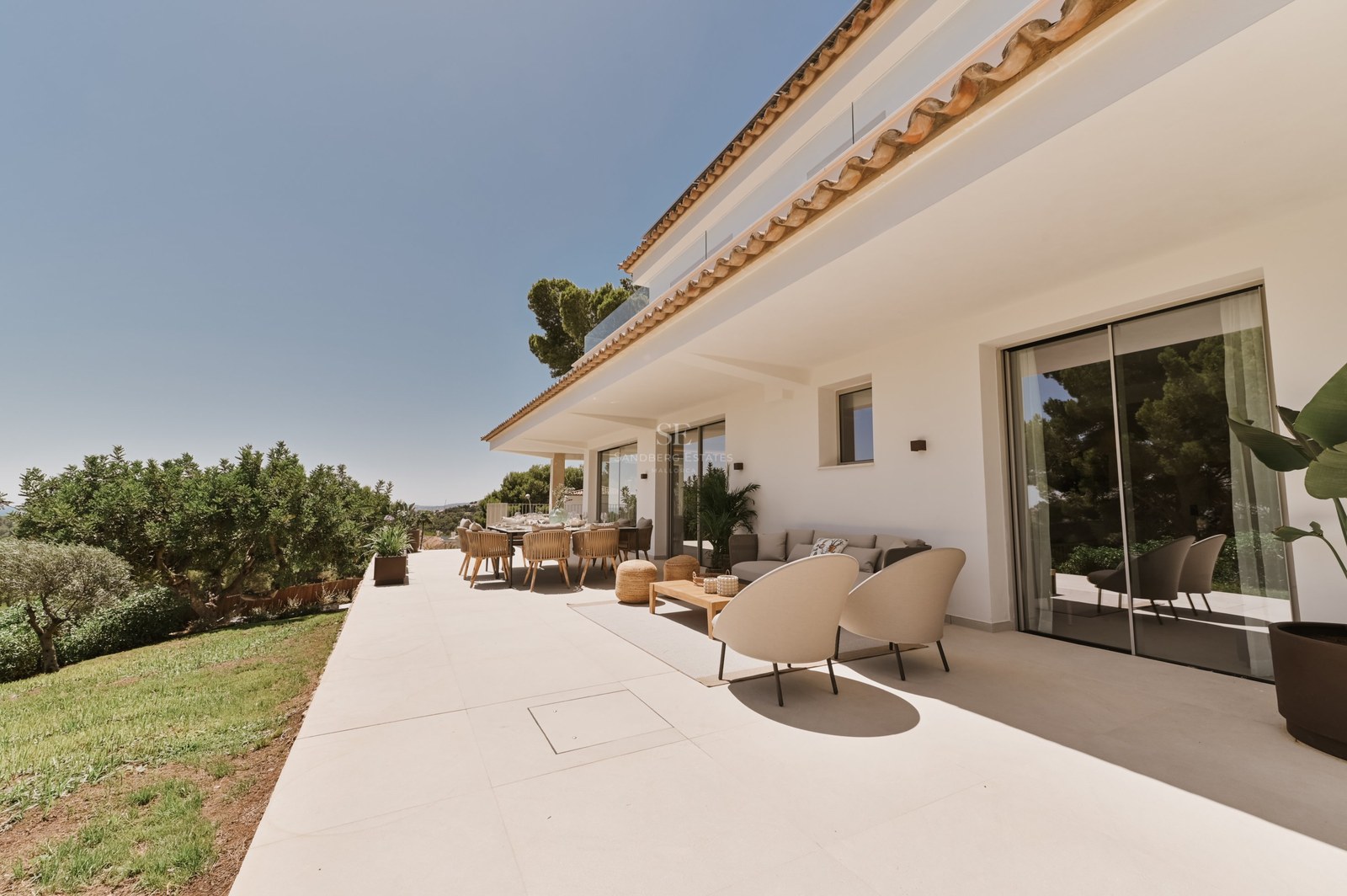 Amplia terraza soleada de piedra con muebles de salón de diseño, zona de comedor y fachada de villa blanca.