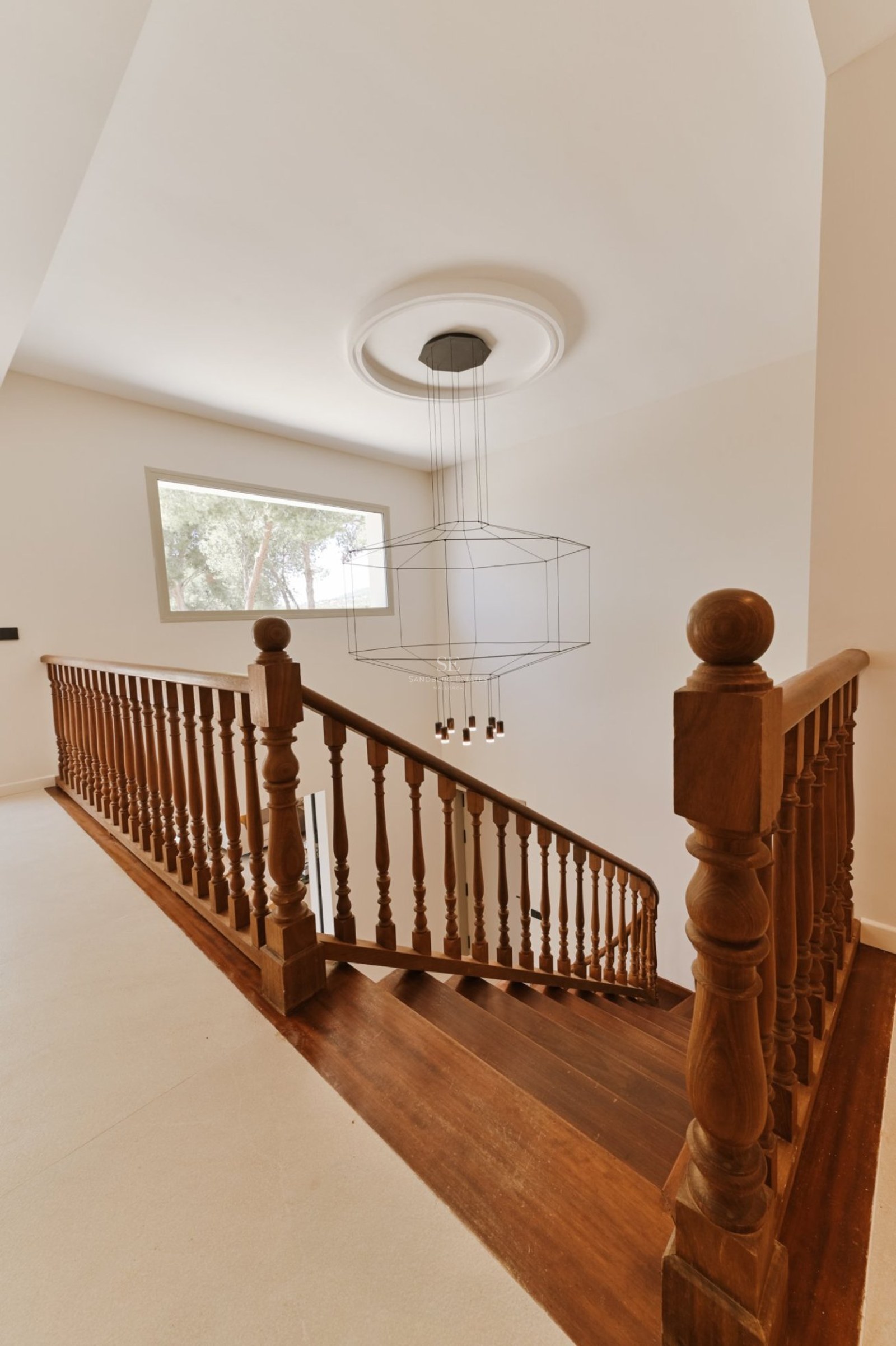 Escalera de madera oscura con barandilla clásica y moderna lámpara de araña de alambre en un pasillo luminoso.