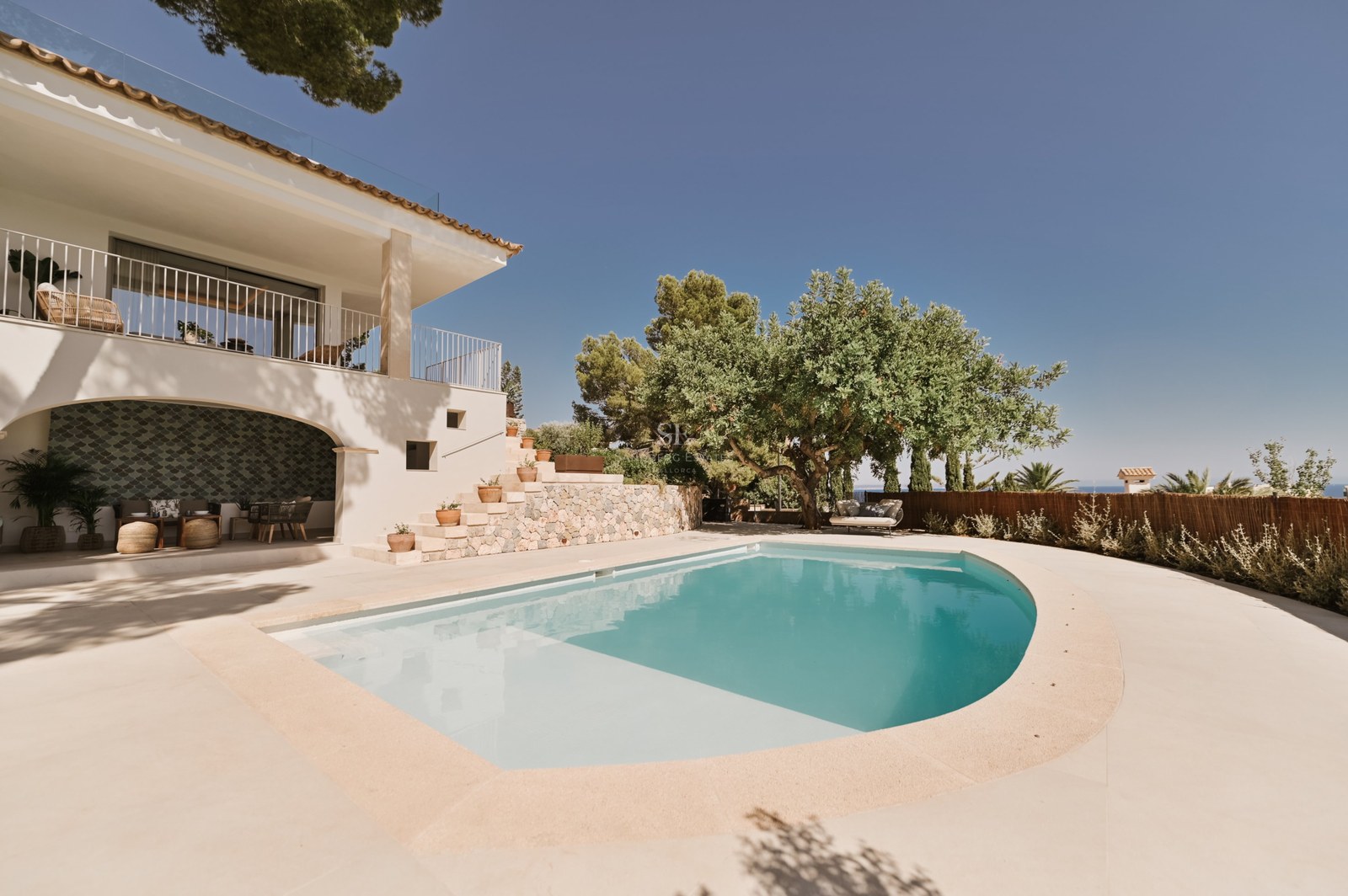 Piscina curva turquesa junto a una villa de paredes blancas con escaleras de piedra y vegetación mediterránea.