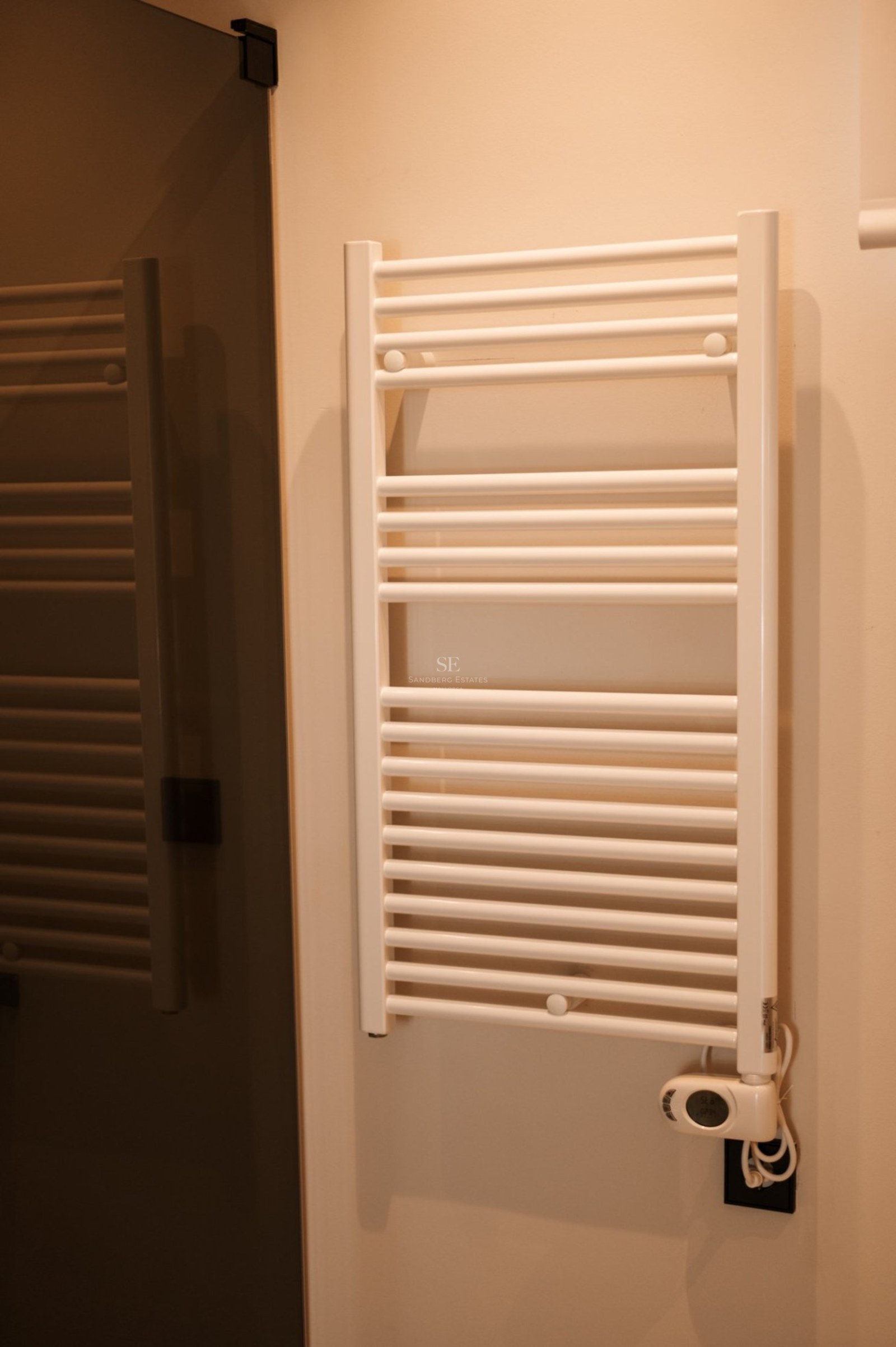 Primer plano de un toallero calefactable blanco de diseño moderno instalado en una pared de tono crema.