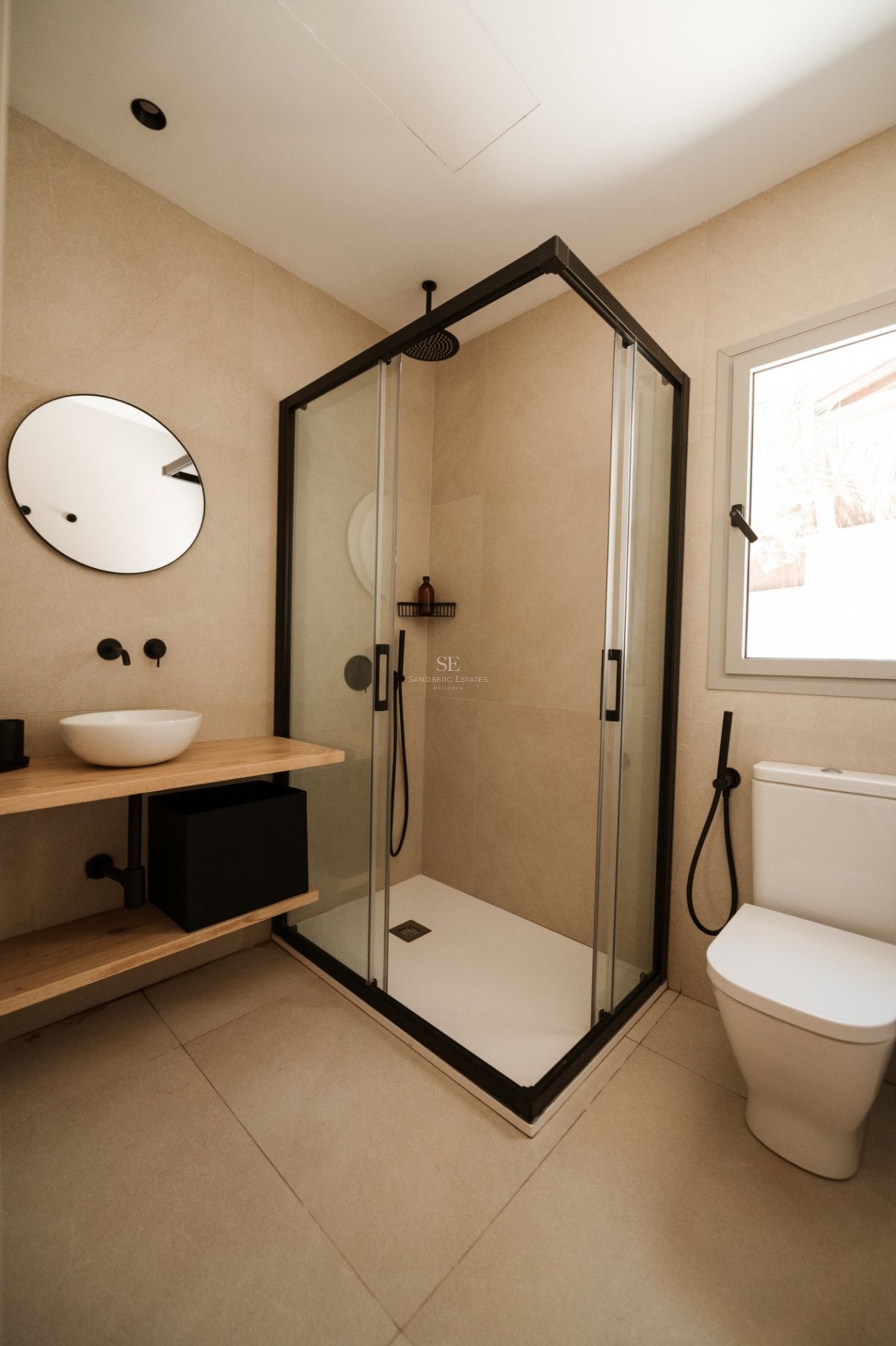 Baño moderno con ducha de cristal, grifería negra mate, encimera de madera y espejo redondo.