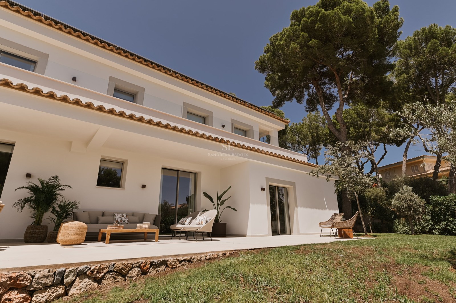 Villa blanca de dos plantas con tejado de terracota, muebles de exterior y pinos bajo un cielo despejado.