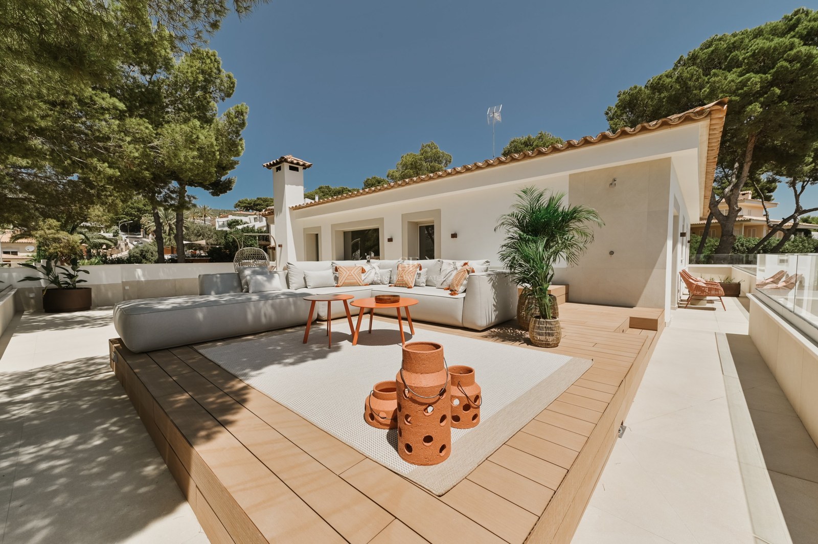 Terraza soleada con sofá en L blanco sobre tarima de madera, linternas de terracota y fachada de villa blanca.