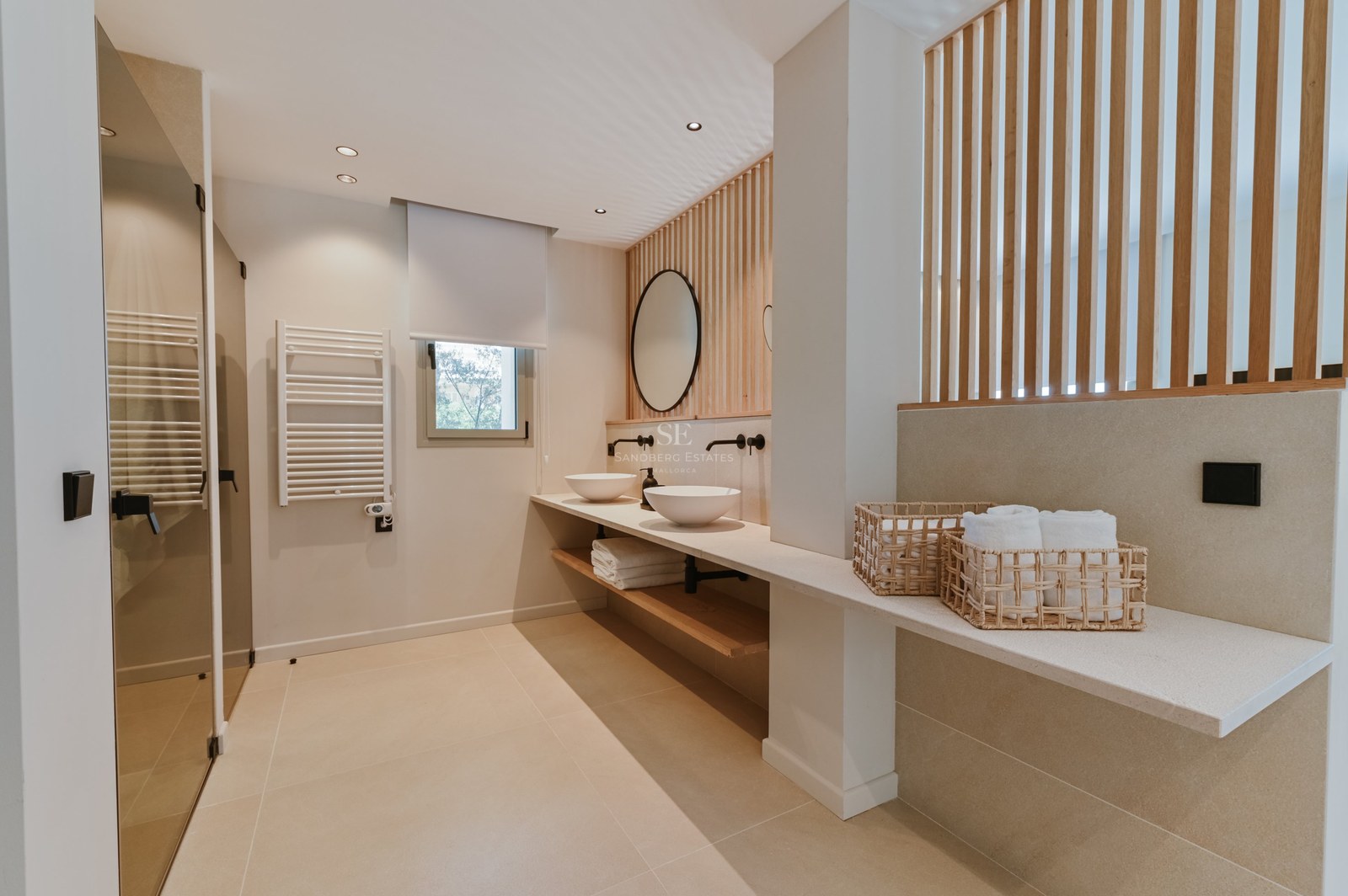 Baño moderno con tocador doble, lavabos sobre encimera, pared de listones de madera y espejo redondo.