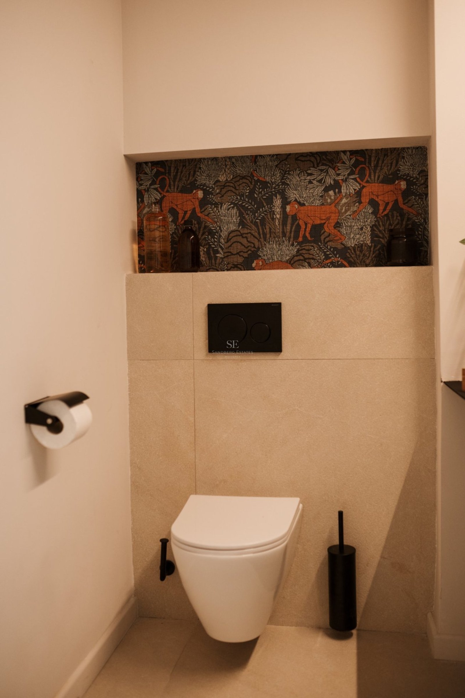 Inodoro suspendido moderno con pulsador negro, azulejos beige y hornacina decorativa con papel pintado de monos.