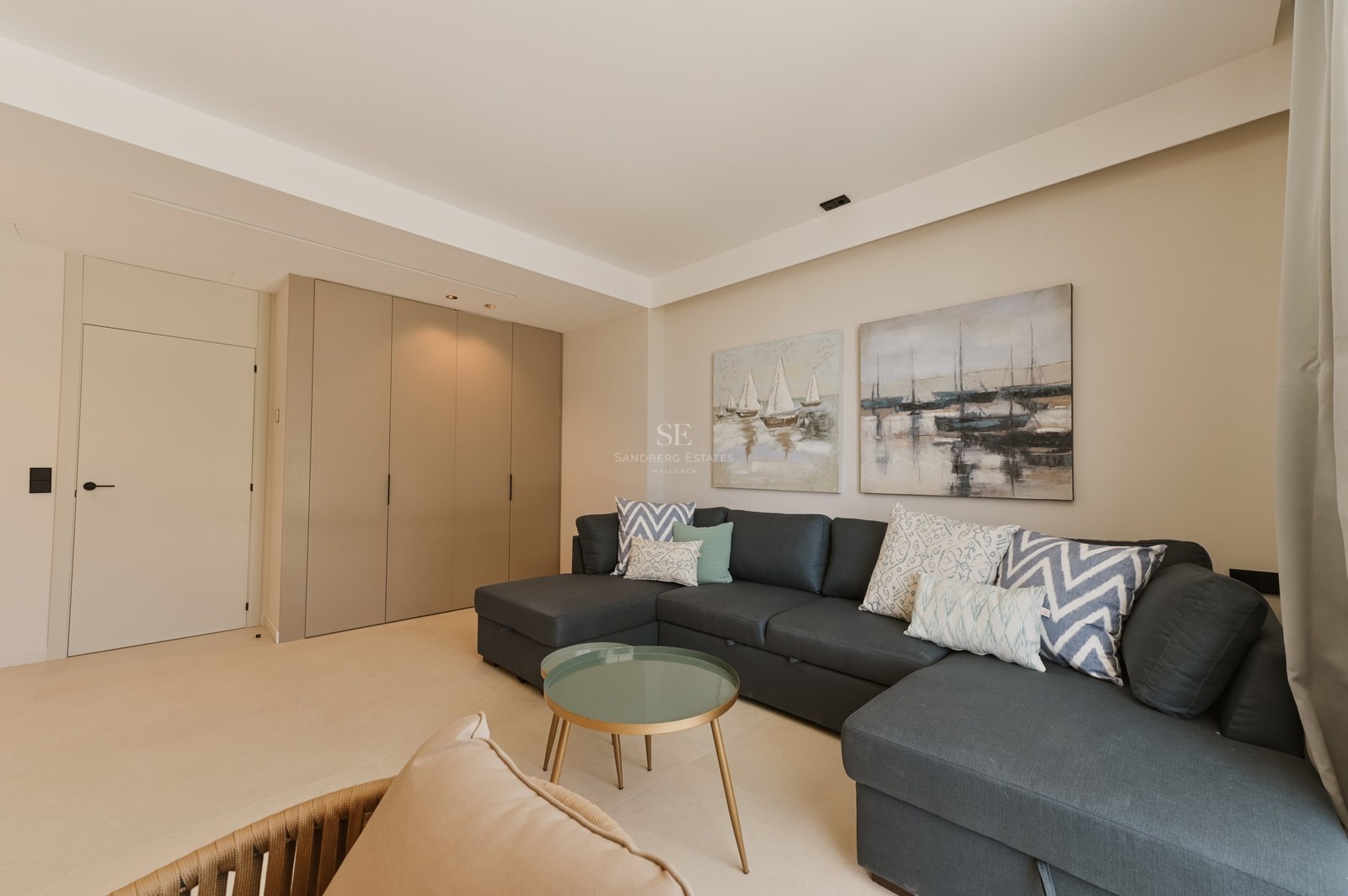 Salón contemporáneo con sofá en L gris oscuro, mesa de centro de cristal y armarios empotrados en tonos neutros.