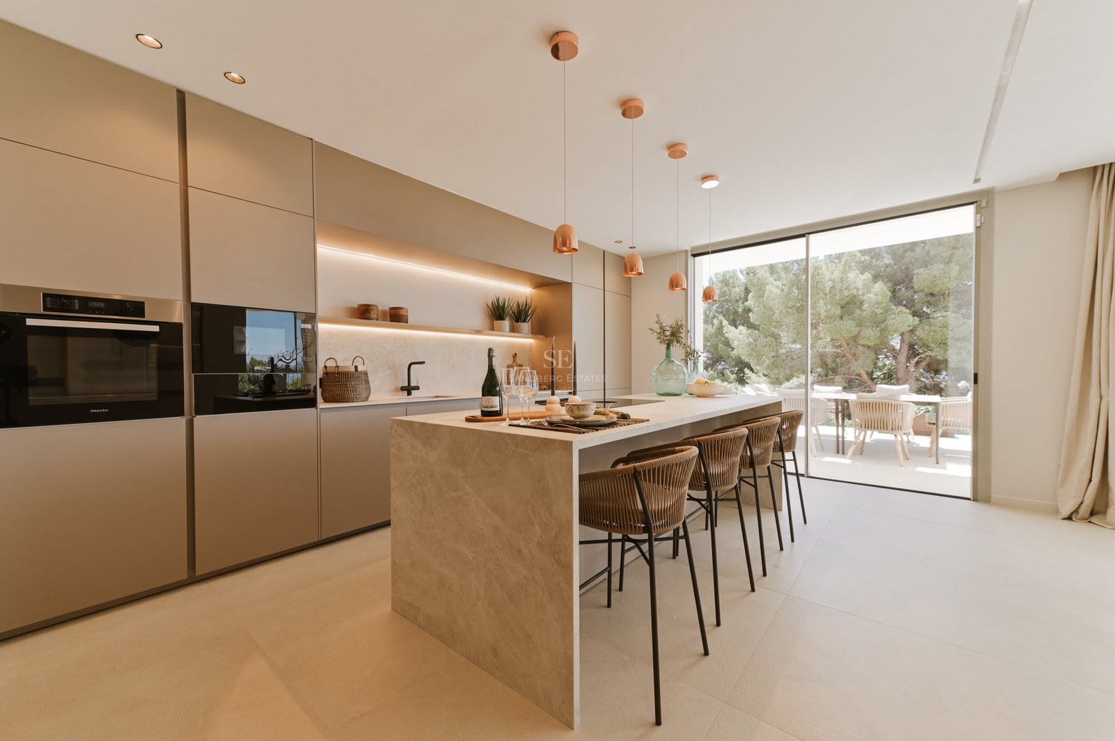 Cocina minimalista moderna con isla de piedra beige, taburetes y ventanales de suelo a techo con vistas al jardín.