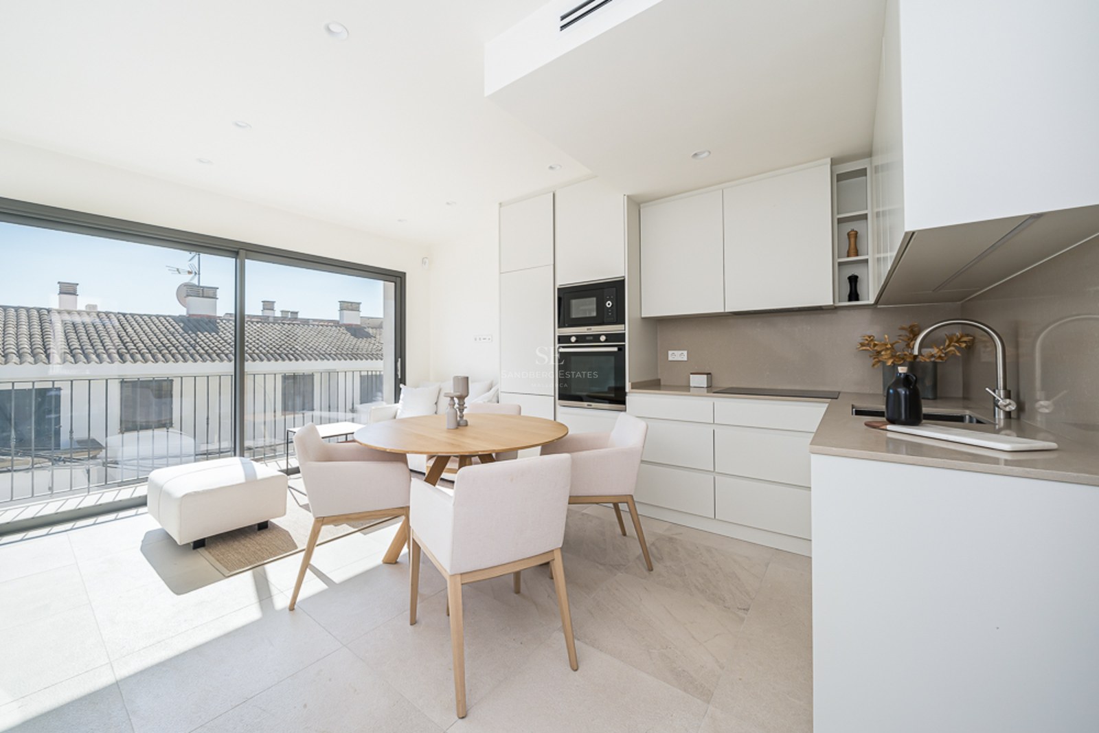 Cocina luminosa con muebles blancos, mesa de comedor de madera y grandes ventanales con vistas a tejados tradicionales.
