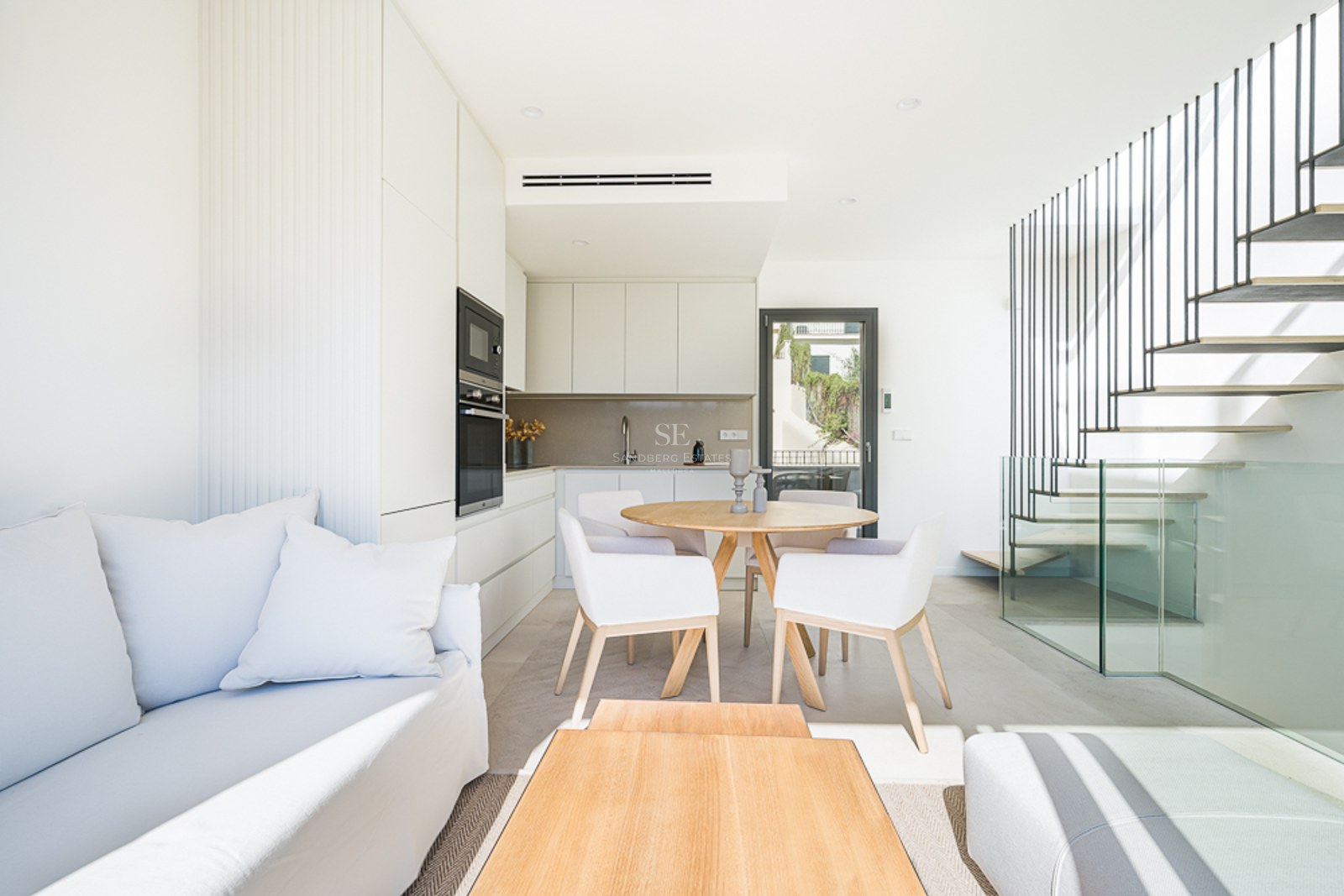 Salón minimalista luminoso con sofá blanco, mesa de comedor de roble, cocina moderna y escalera flotante con cristal.