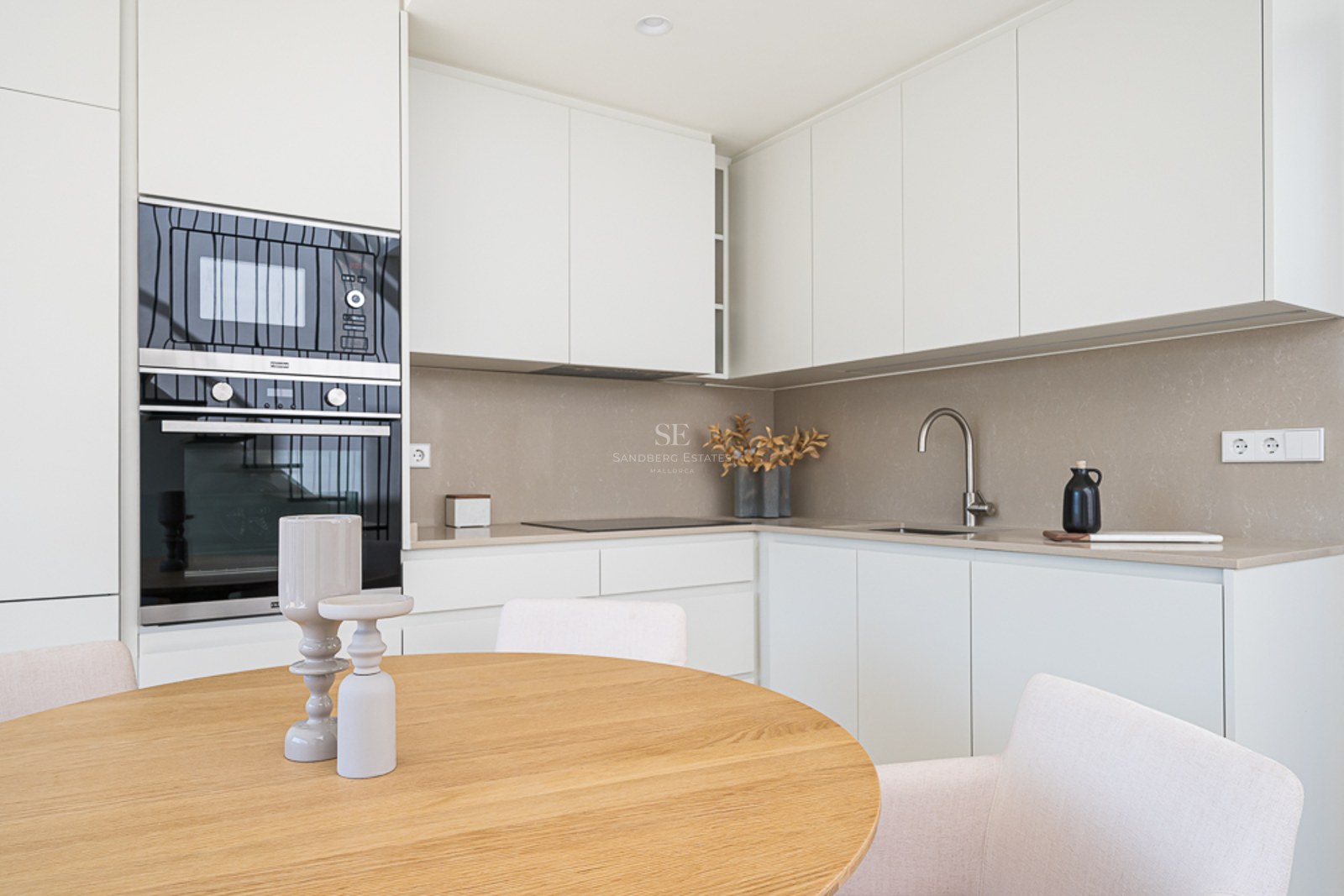 Cocina blanca moderna con encimera de cuarzo, electrodomésticos integrados y mesa de comedor redonda de roble.