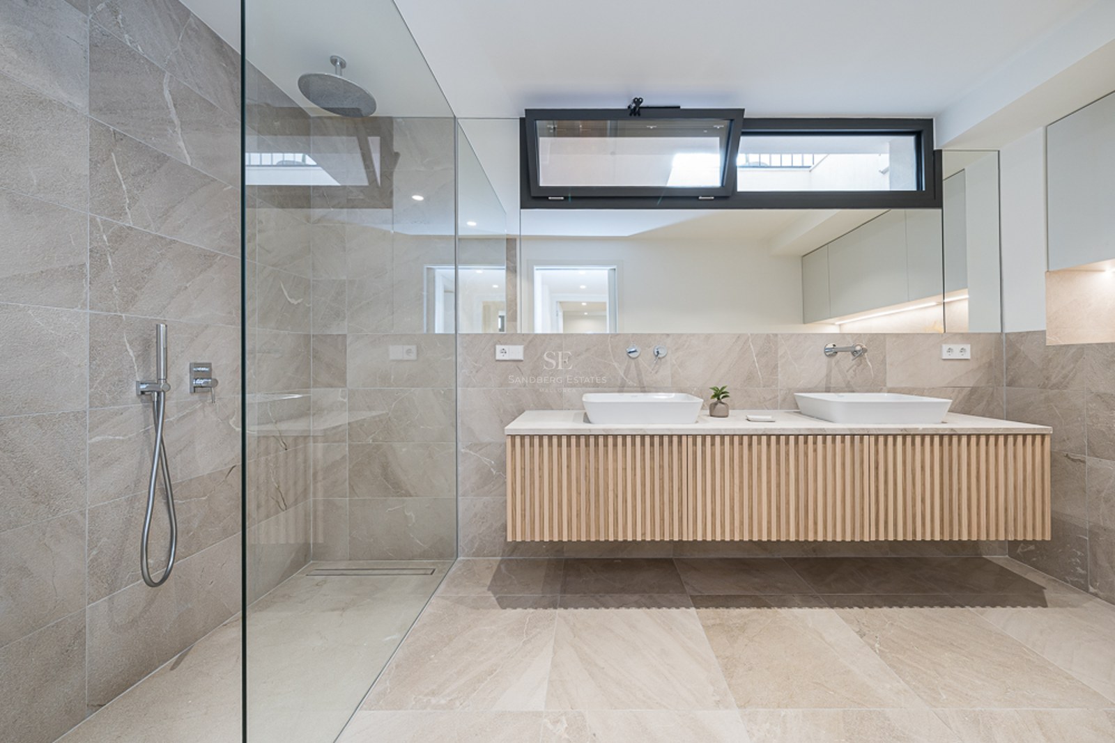 Baño moderno con ducha de cristal, azulejos de piedra color arena y mueble doble suspendido de madera.