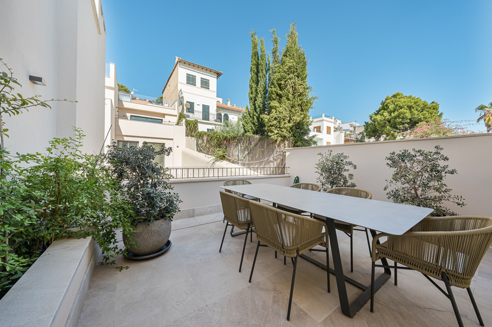 Terraza exterior contemporánea con mesa de comedor grande y seis sillas de cuerda trenzada sobre suelo de piedra clara.