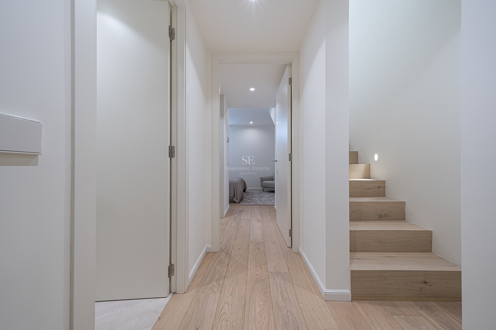 Pasillo moderno con suelos de madera clara, paredes blancas y escalera de madera con iluminación integrada.