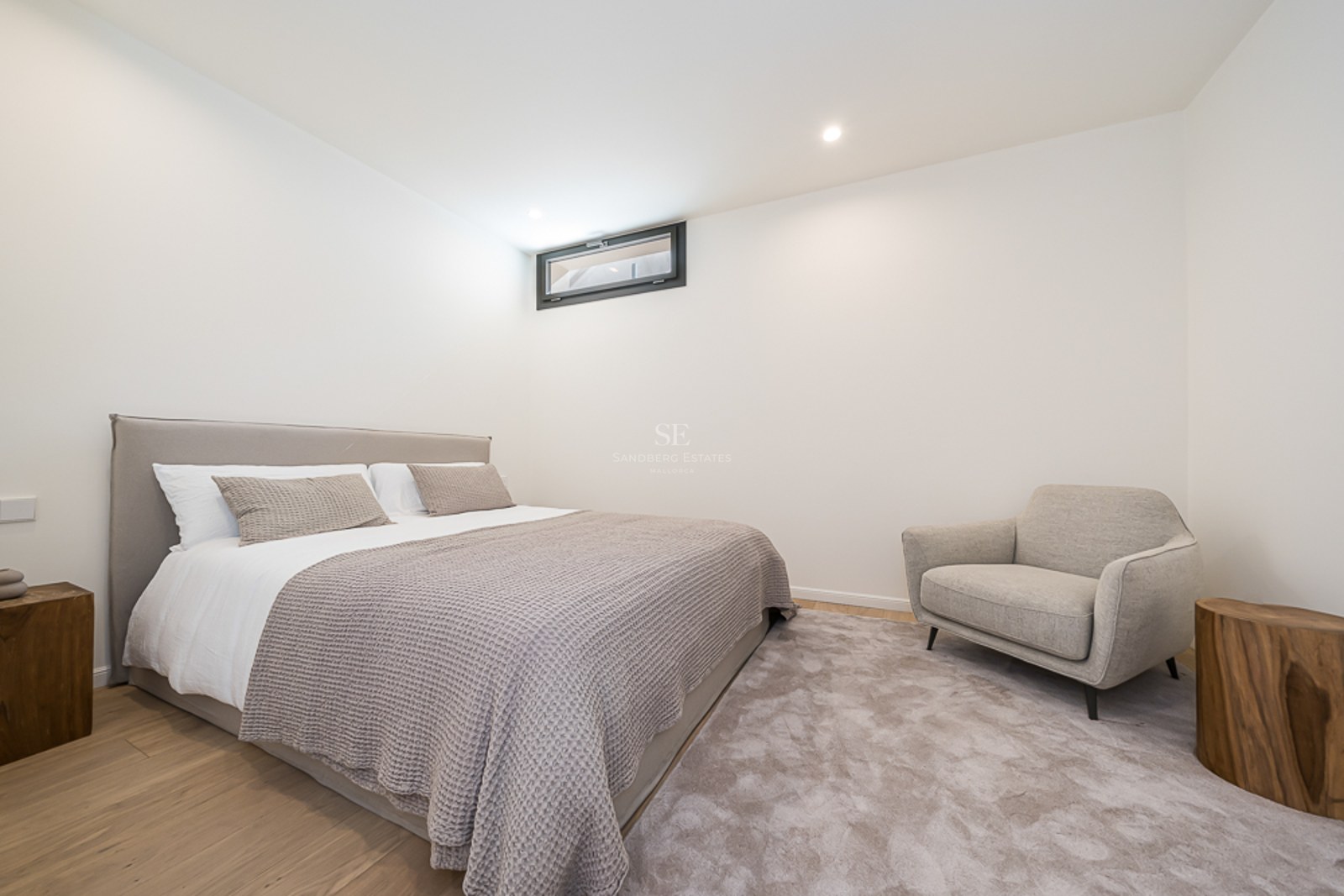 Dormitorio moderno con cama tapizada, mesitas de madera natural, sillón gris y suelos de roble claro.