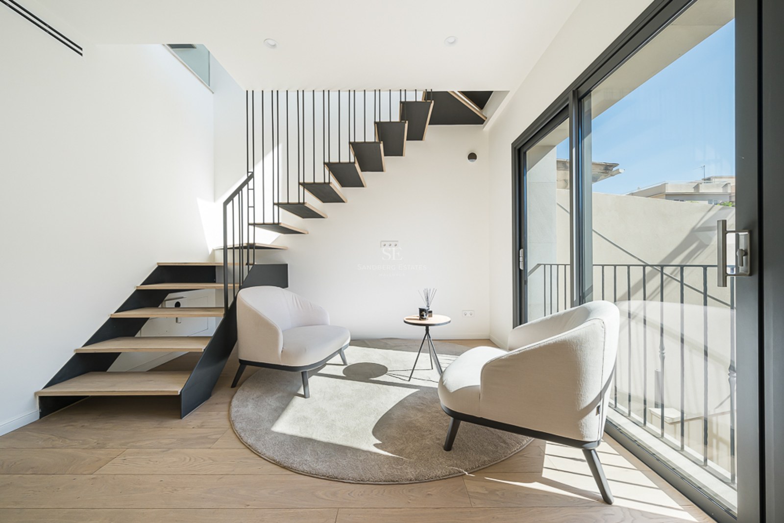 Espacio moderno con escalera flotante de madera, dos sillones crema y ventanales de suelo a techo.