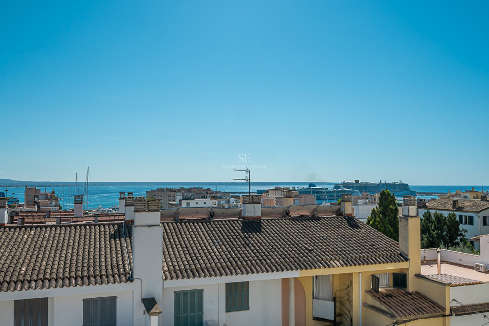 Vista elevada sobre tejados tradicionales hacia el mar azul con un crucero y veleros en el puerto.