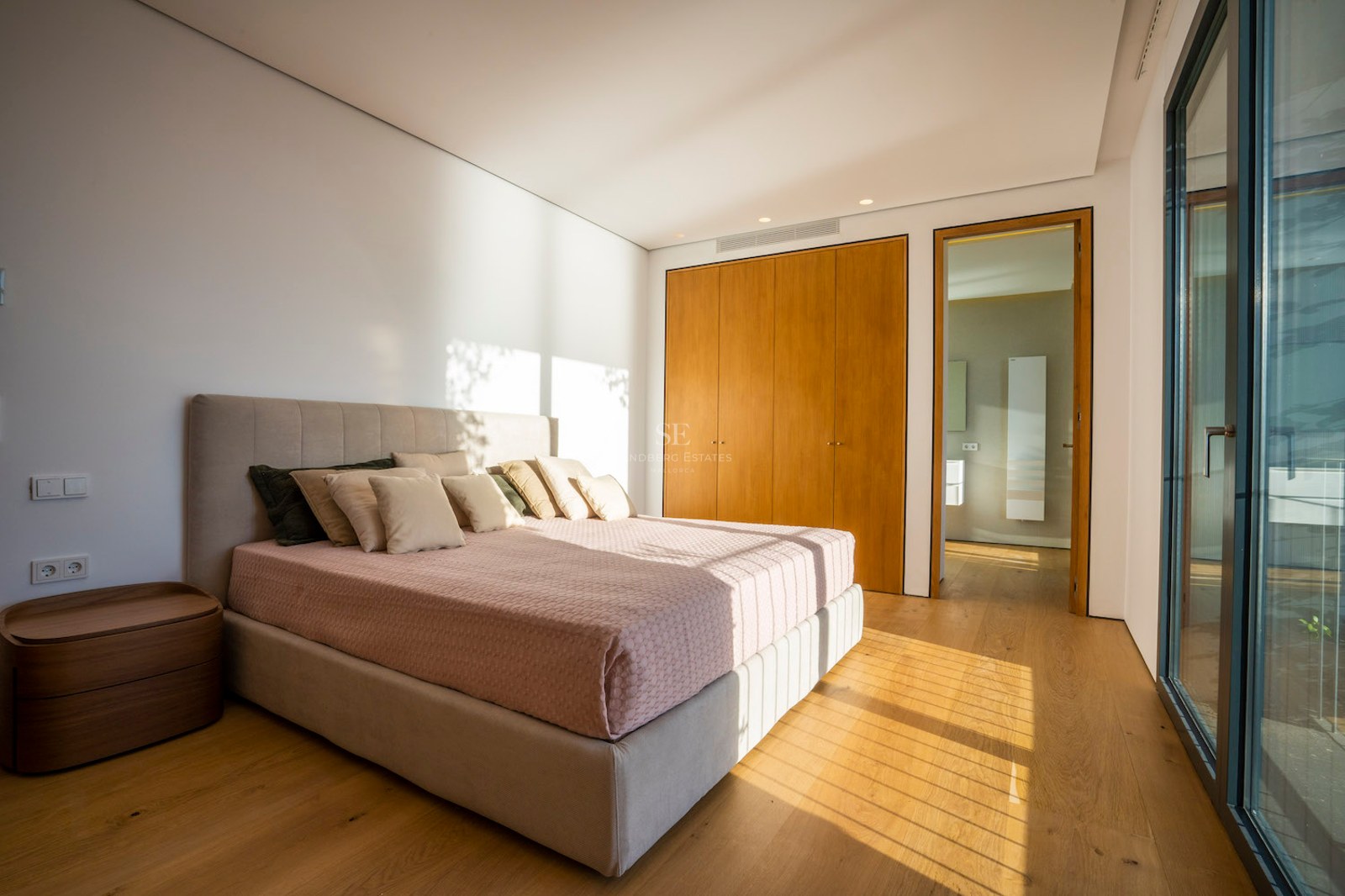 Modern bedroom with oak floors, built-in wooden wardrobes, a large bed, and floor-to-ceiling glass doors.