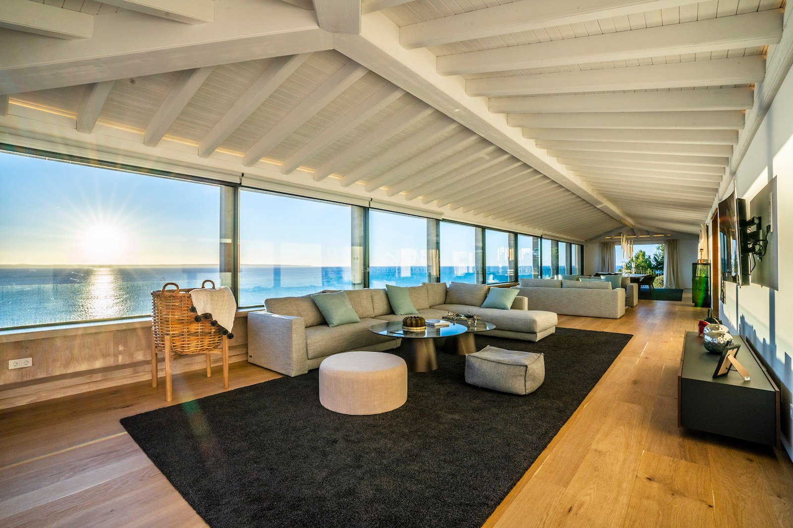 Modern living room with white beamed ceilings, oak floors, and floor-to-ceiling windows overlooking the ocean at sunset.