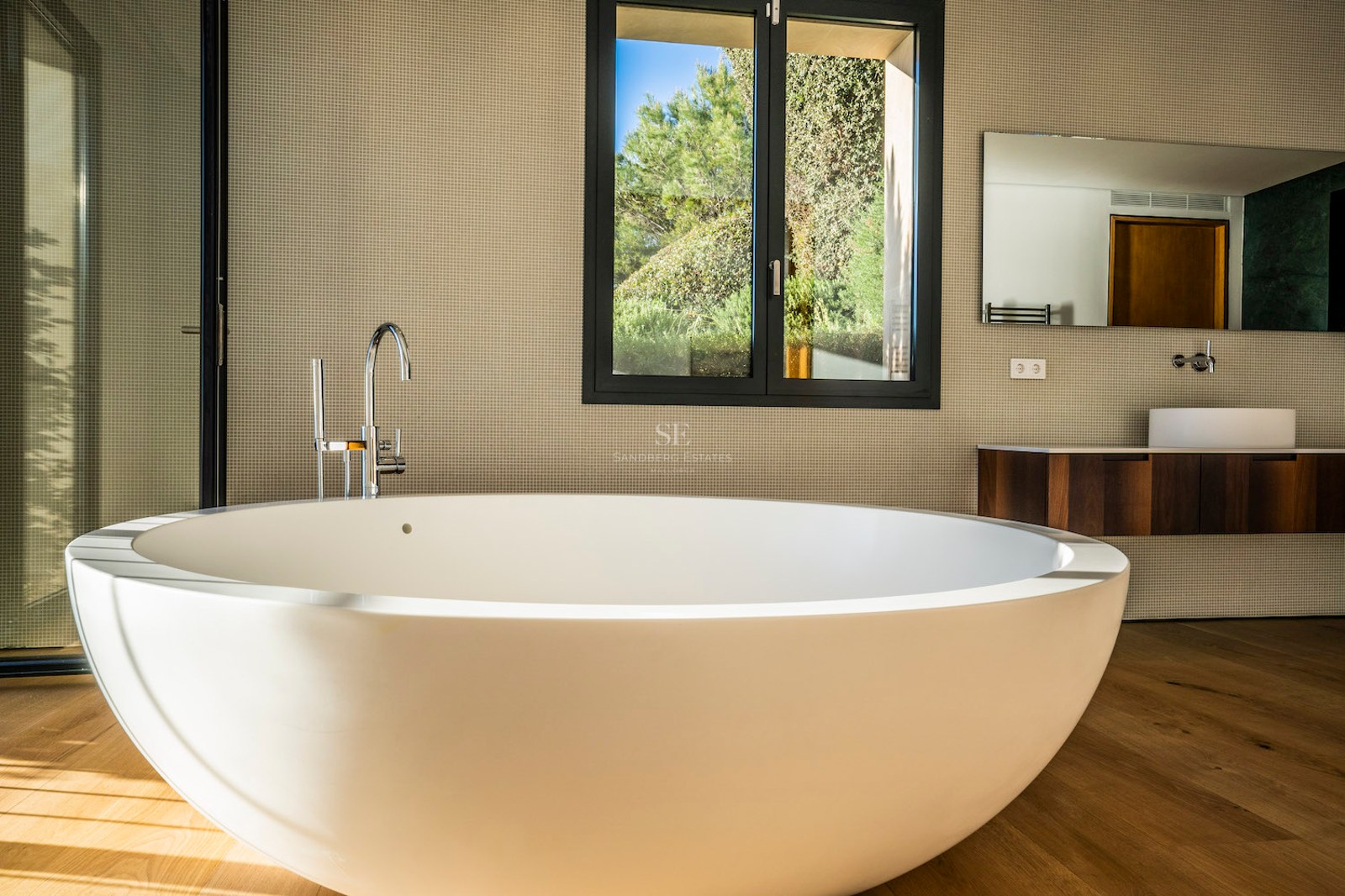 Modern master bathroom featuring a large white oval freestanding tub on oak floors with garden views through a window.