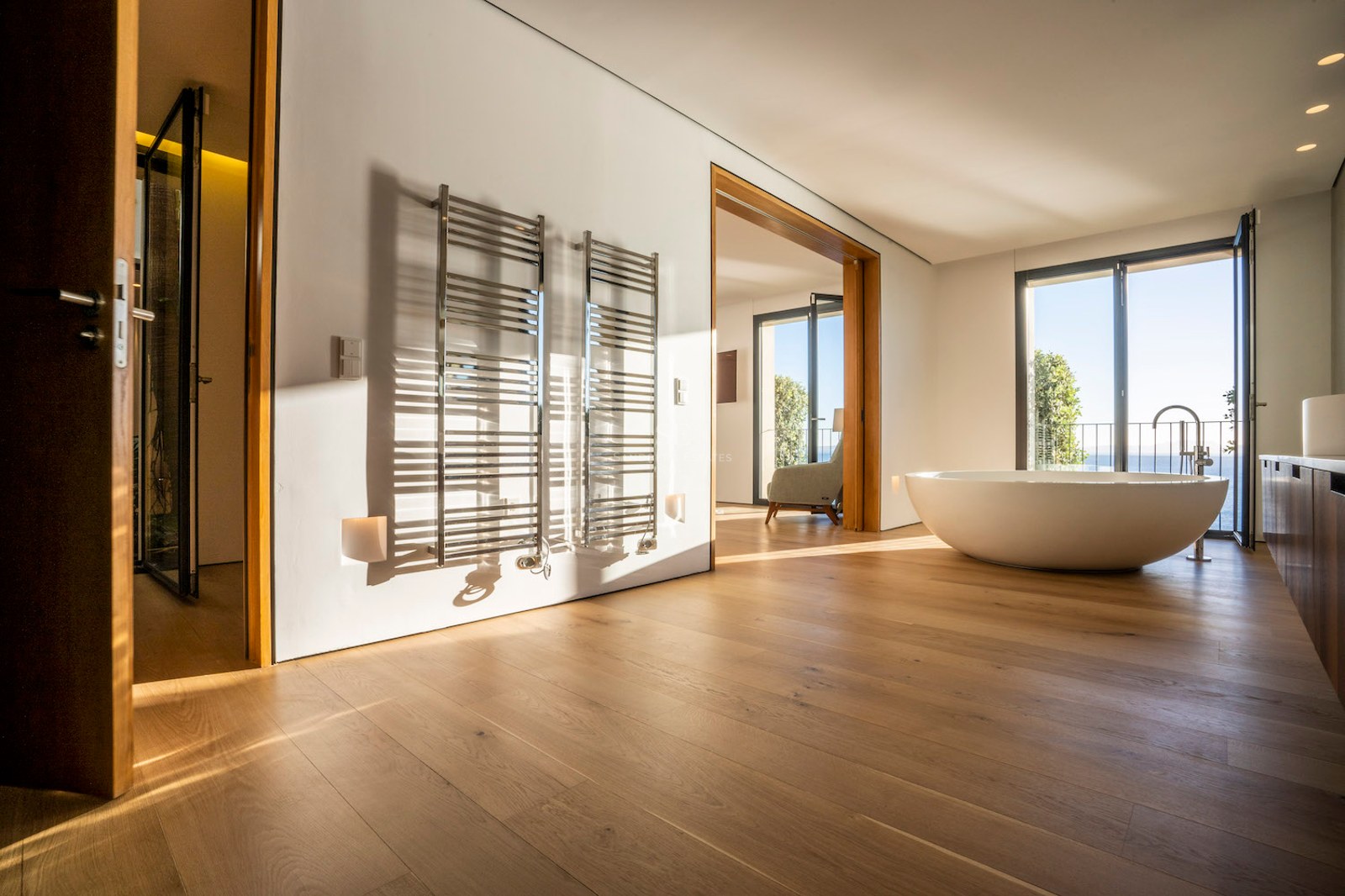 Minimalist master bathroom with oak floors, oval freestanding tub, chrome towel rails, and Mediterranean sea views.