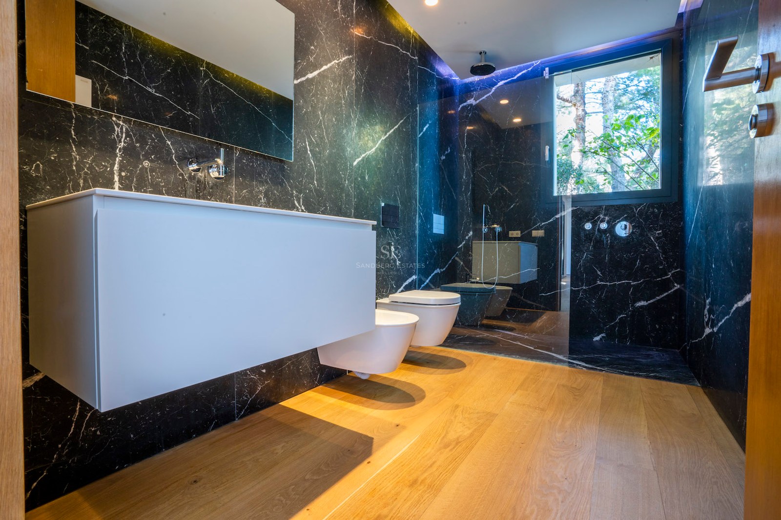 Modern bathroom with black veined marble walls, oak flooring, floating white vanity, and walk-in glass shower.