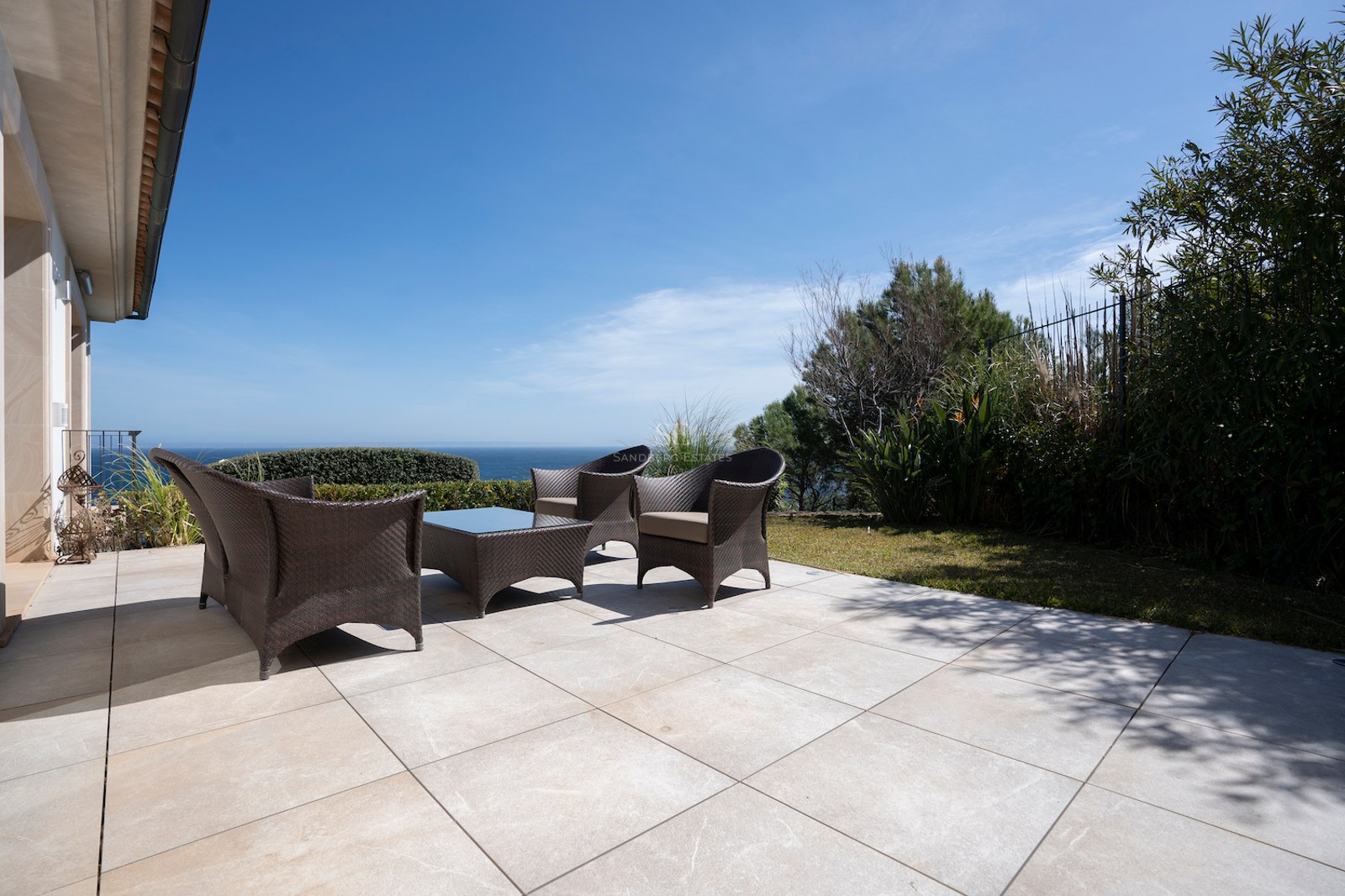 Large stone tile terrace with dark wicker lounge furniture overlooking the ocean under a clear blue sky.