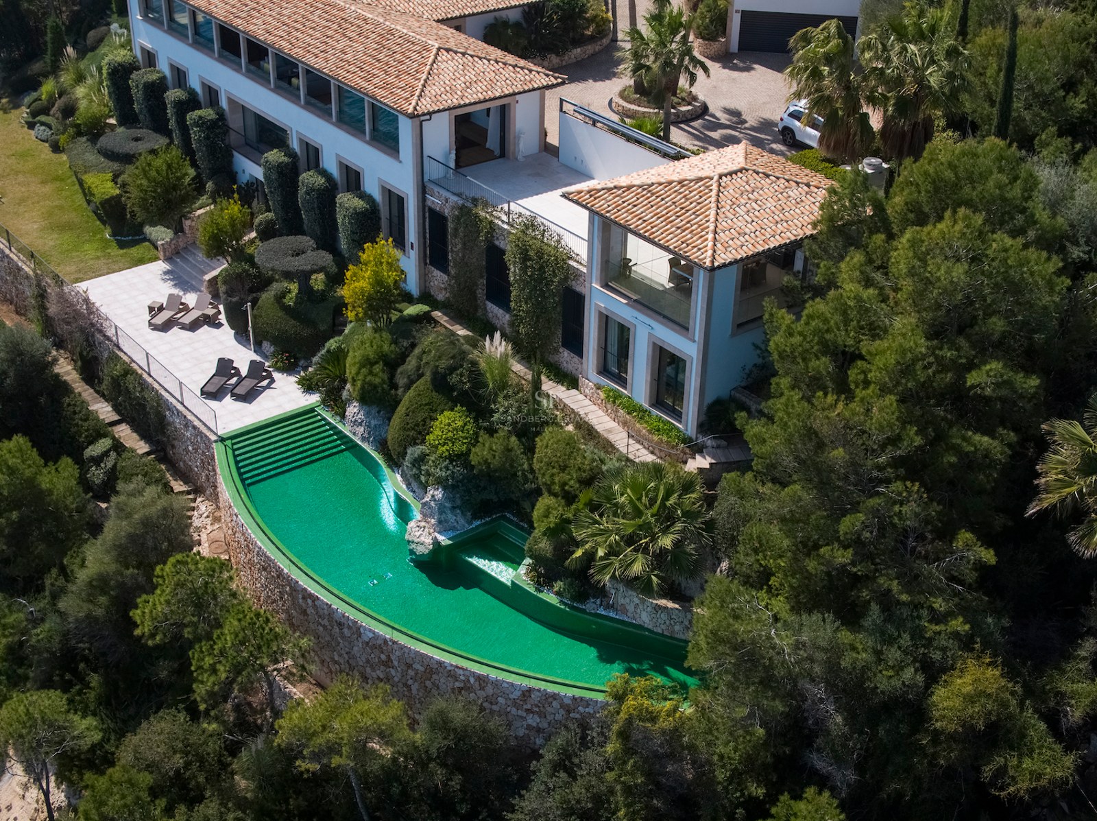 Aerial view of a luxury villa featuring a curved emerald green infinity pool, stone walls, and terracotta roof.