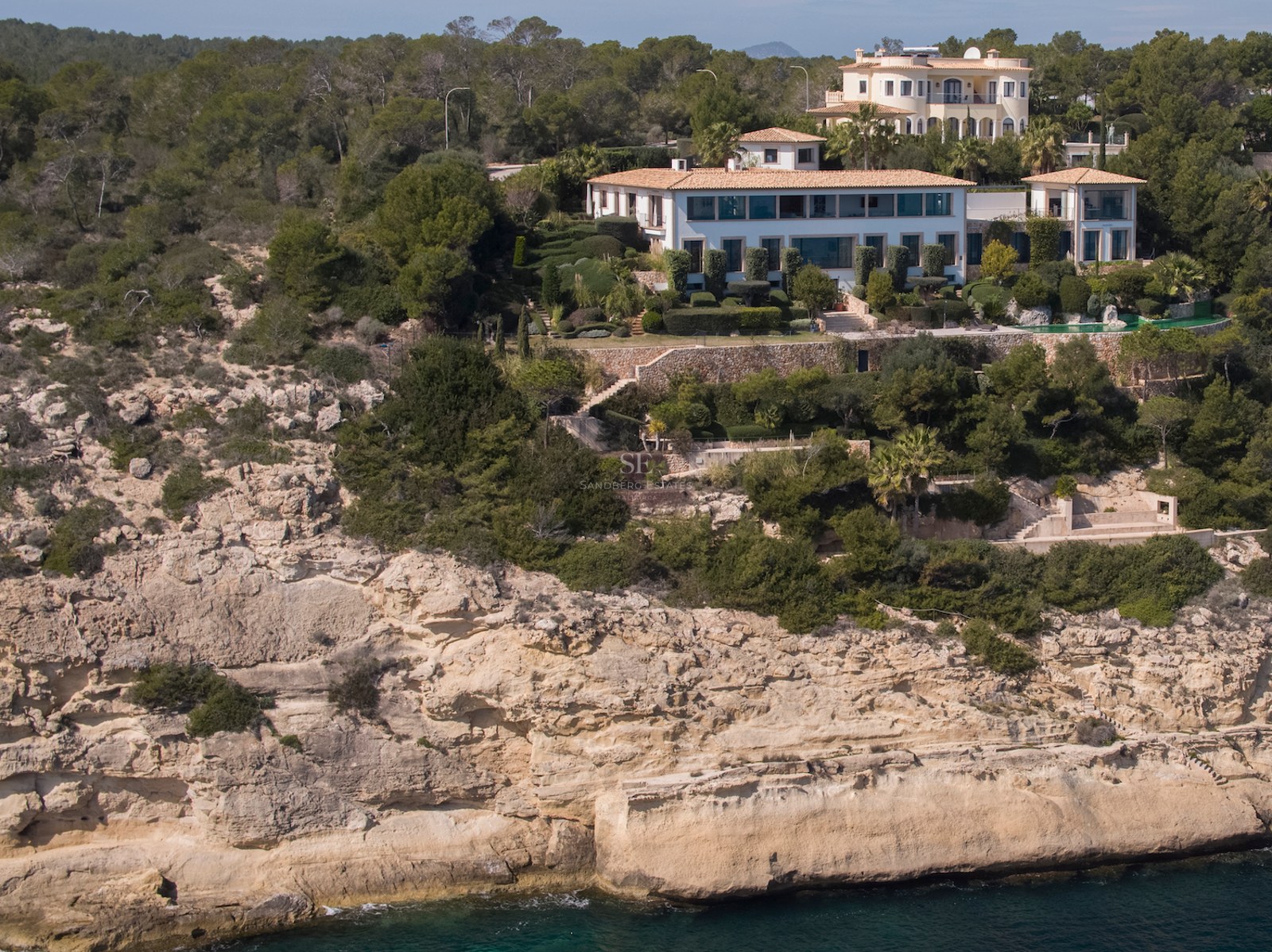 An aerial view of a large white Mediterranean villa perched on a rocky limestone cliff overlooking the blue ocean.