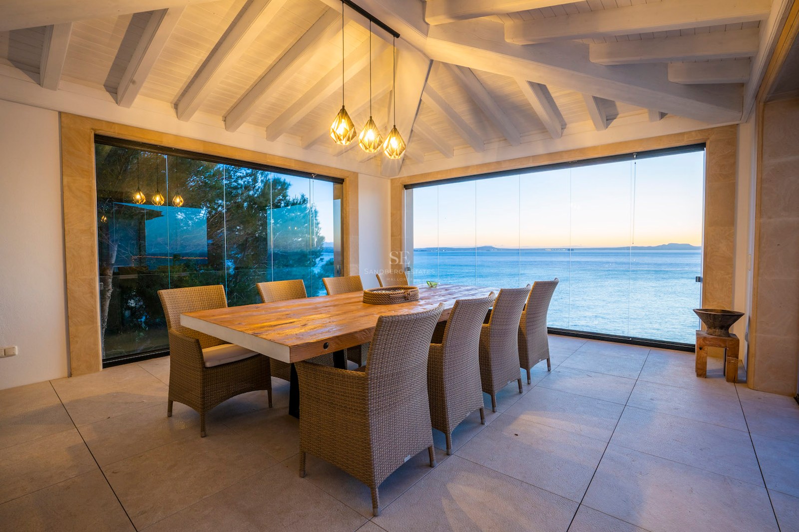 Large wooden dining table on a terrace with glass walls overlooking the blue sea at dusk under a beamed ceiling.