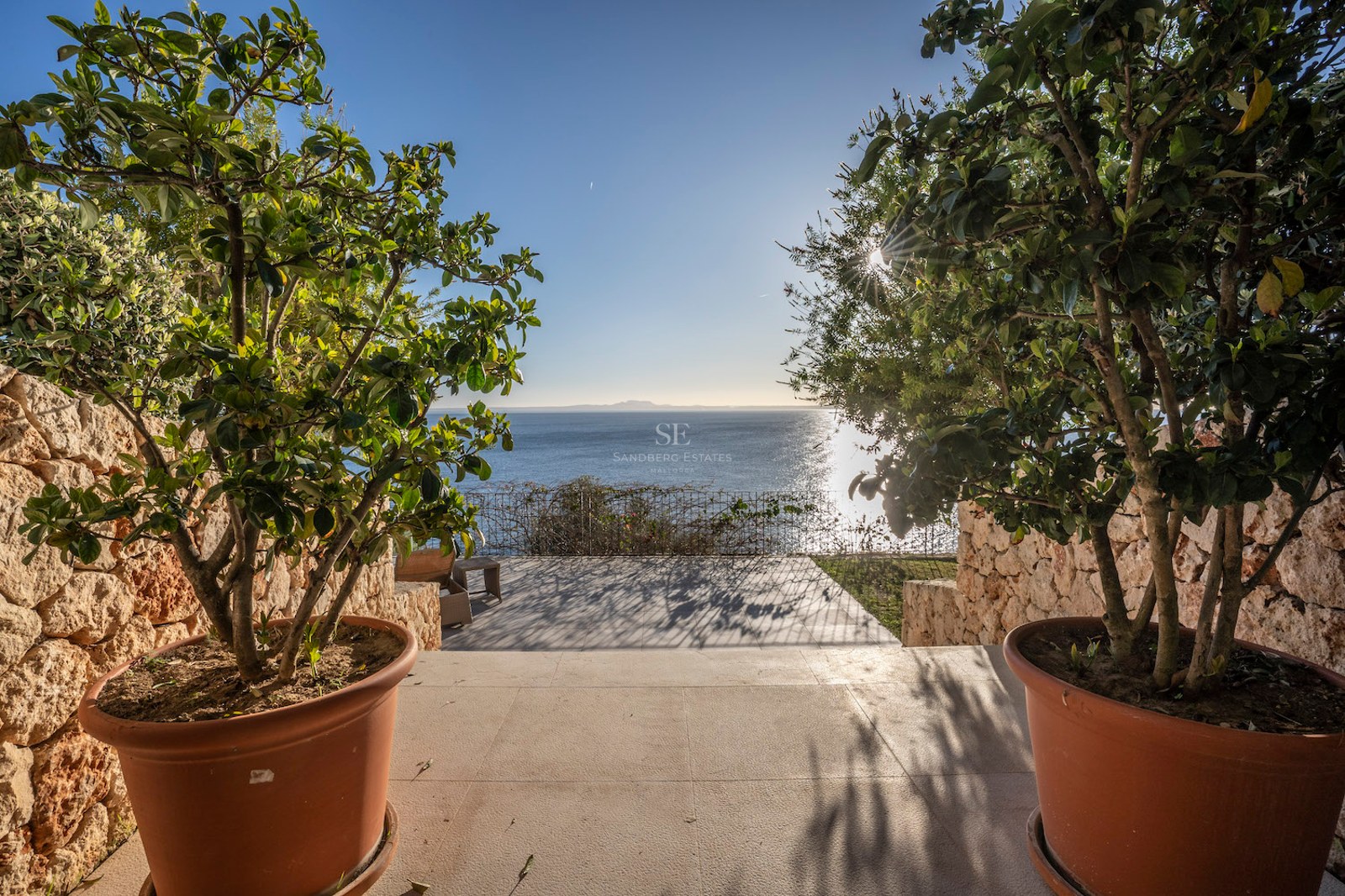 View of the blue sea from a stone terrace framed by potted plants and dry stone walls under a clear sky.