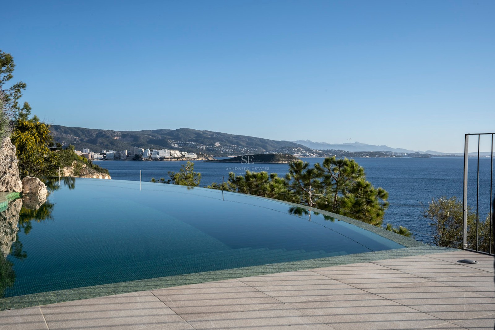 An infinity swimming pool with clear blue water overlooking the sea and a mountainous coastline under a clear sky.