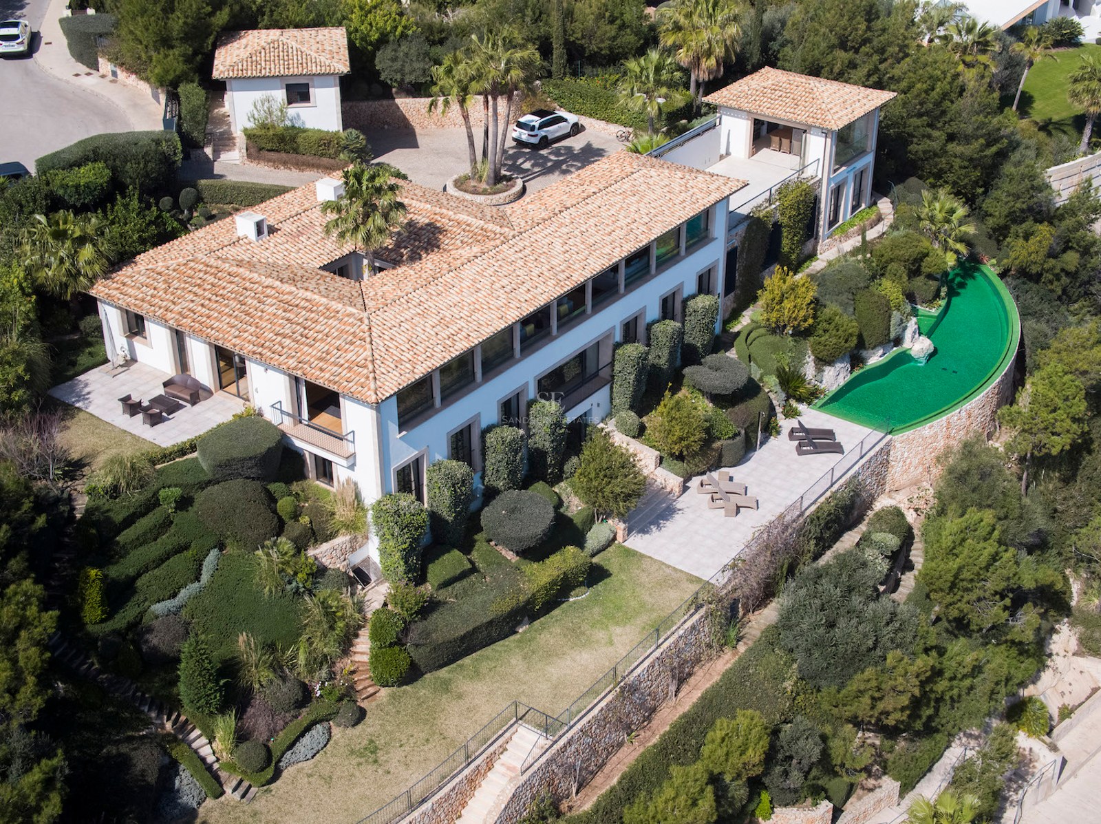High-angle drone view of a large white villa with a terracotta roof, curved pool, and lush manicured gardens.