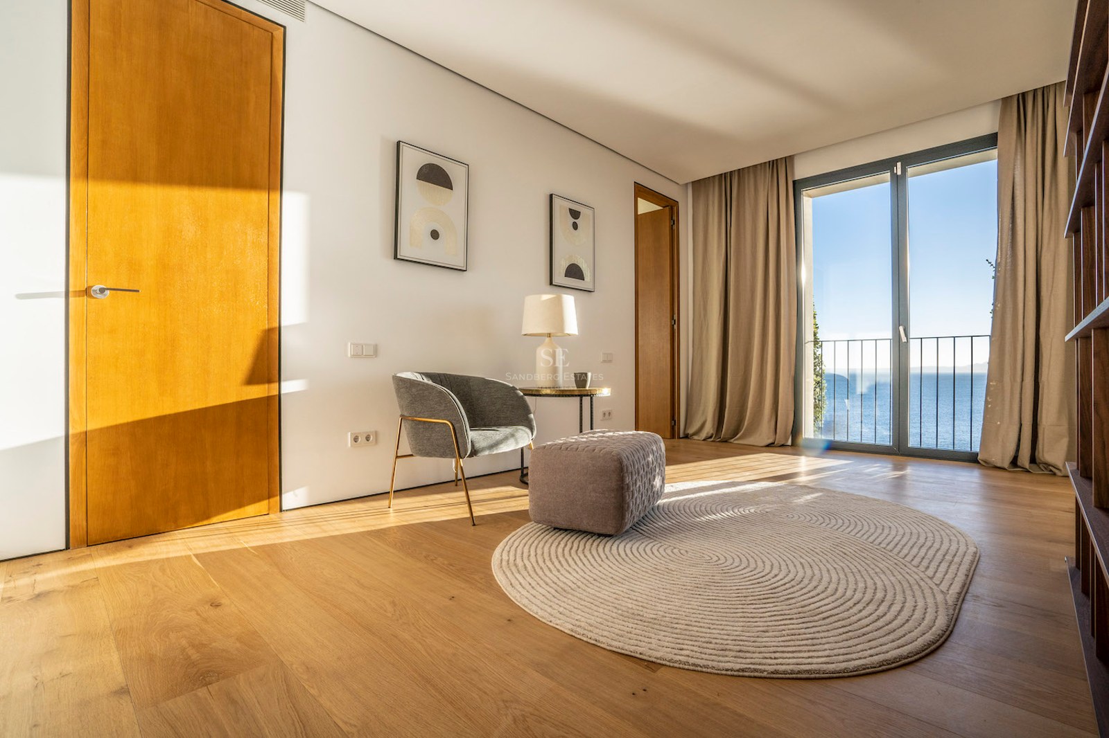 Modern room with oak floors, grey armchair, and glass doors opening to a Mediterranean sea view.