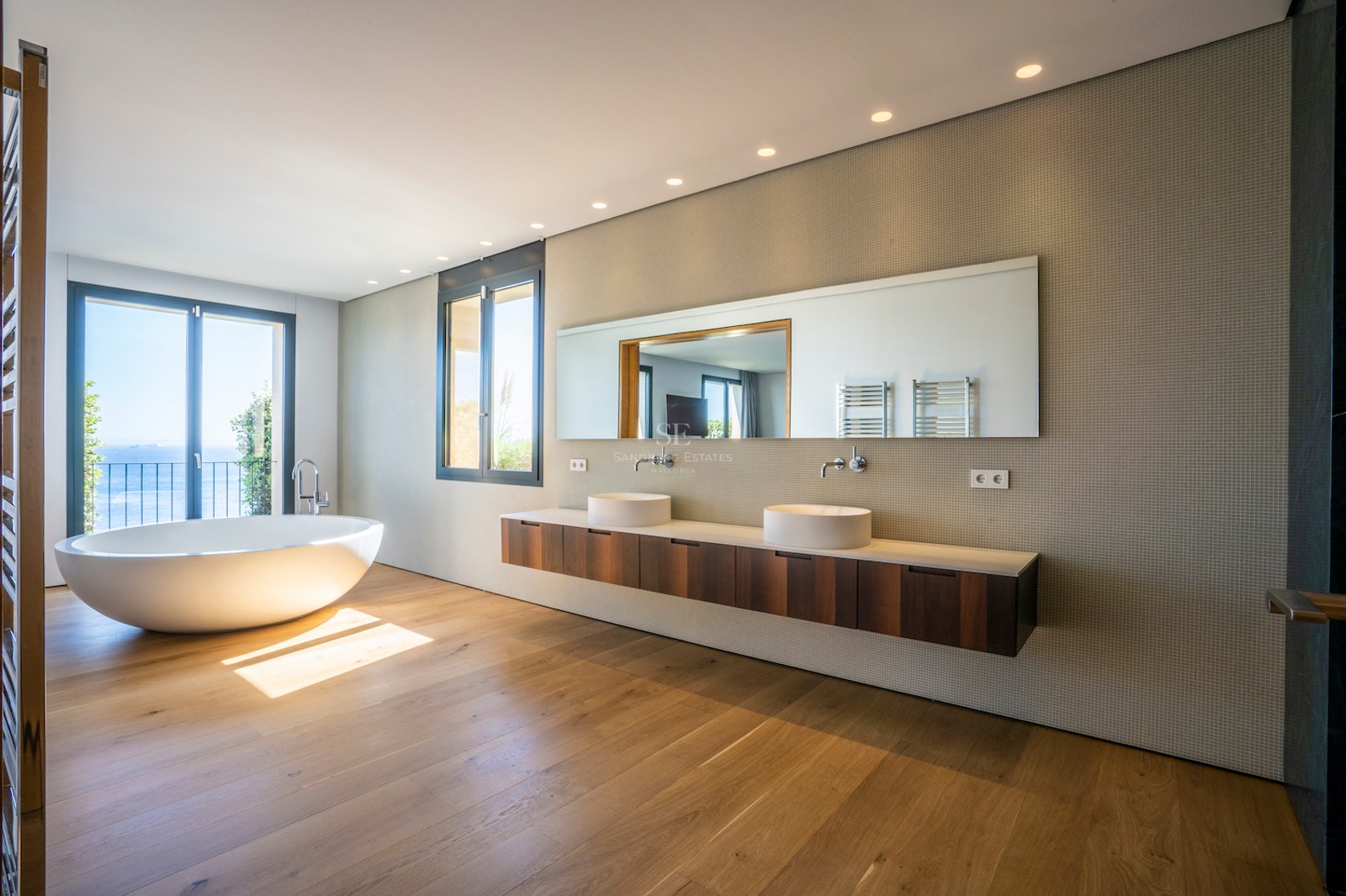 Modern bathroom with freestanding oval bathtub, oak floors, double vanity, and sea views through a glass door.