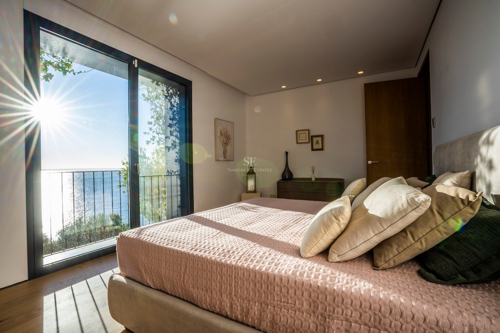 Bright bedroom with a large bed, pink linens, oak floors, and a floor-to-ceiling window overlooking the blue sea.