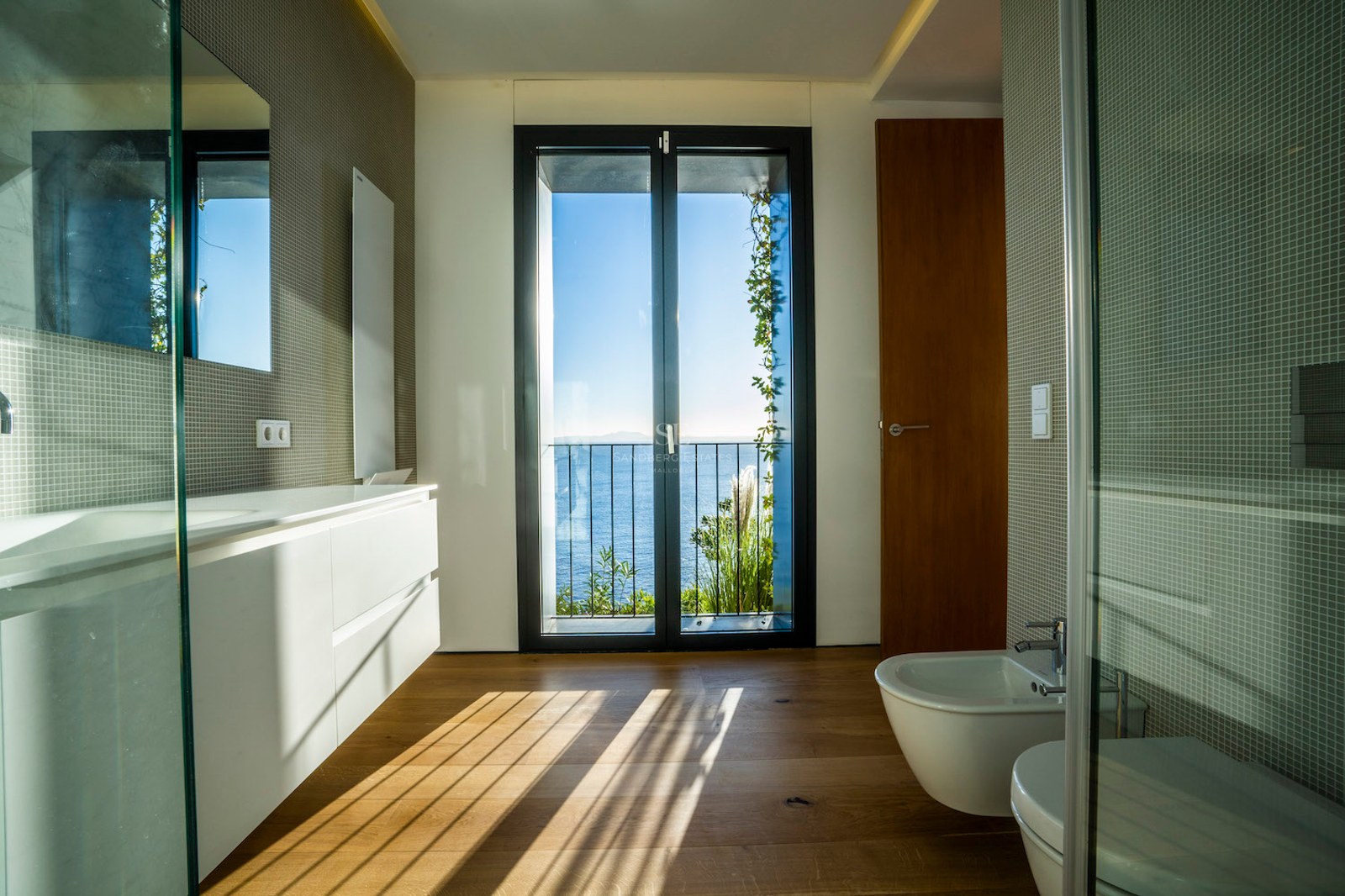 Modern bathroom with oak floors, white vanity, and glass door opening to a balcony with sea views.