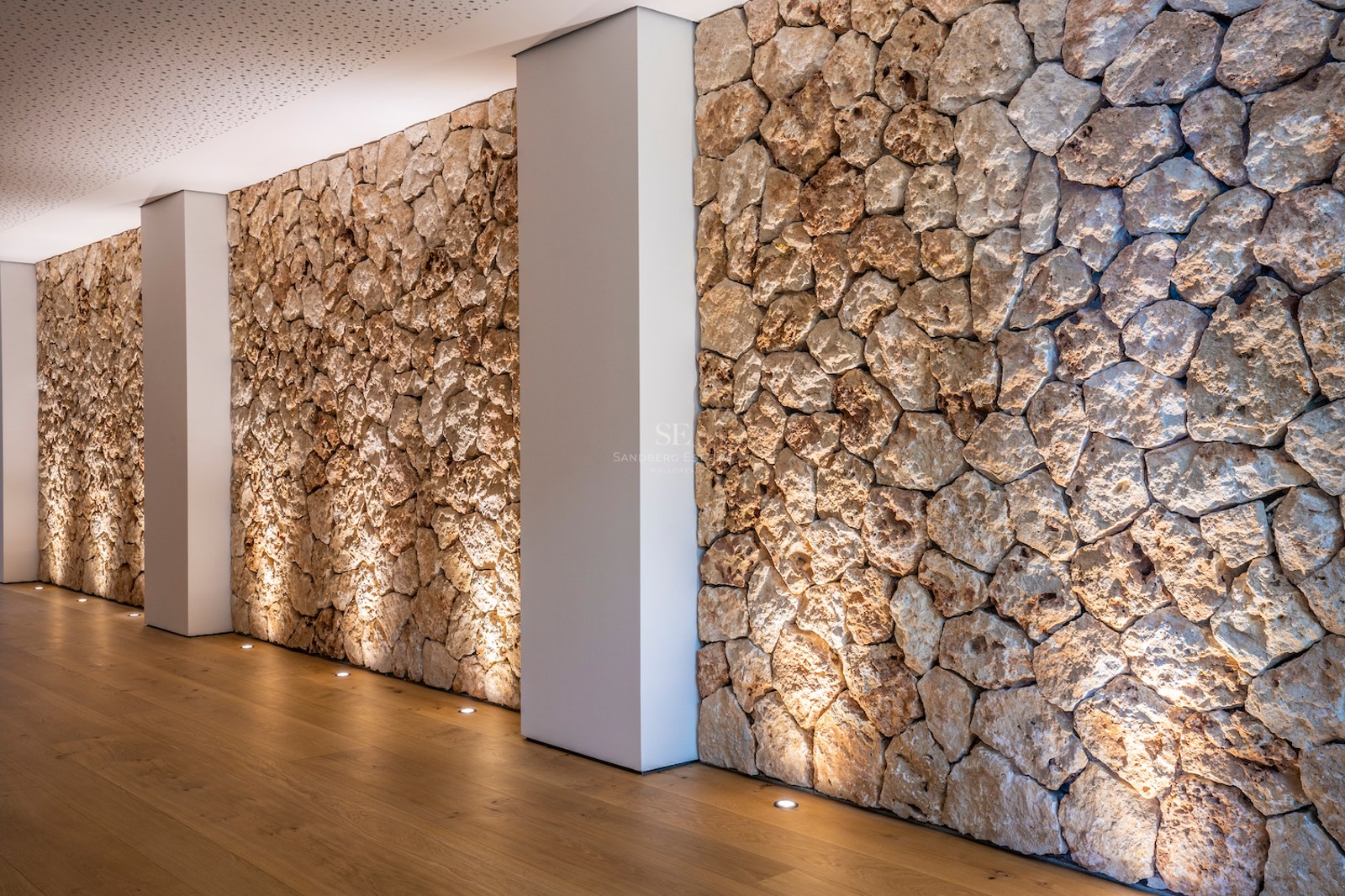 Interior corridor featuring a rustic dry-stone wall illuminated by floor uplights and light oak flooring.
